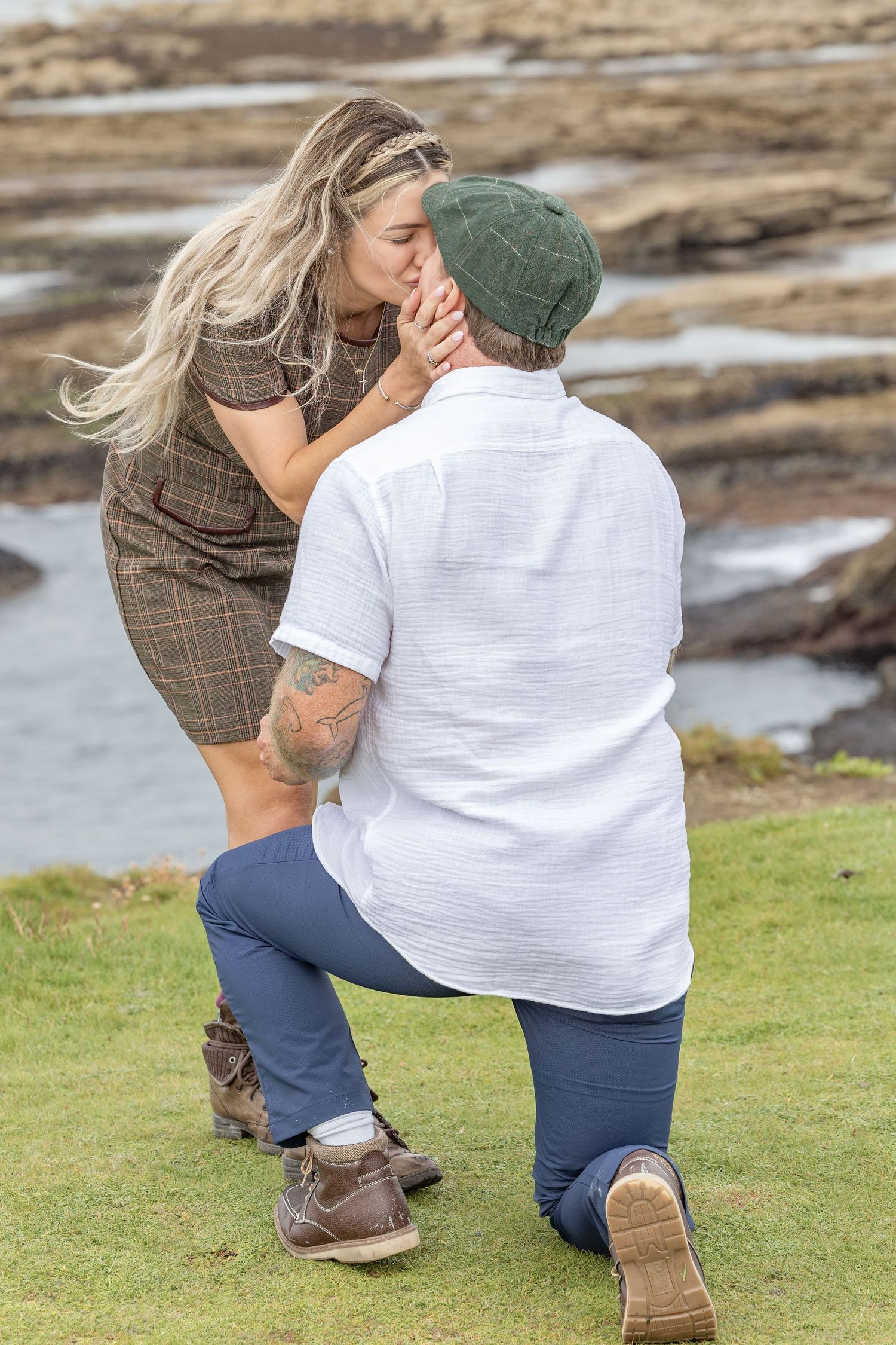 Man on one knee proposing to blonde woman near rocky coastal shoreline while she leans in to kiss him.