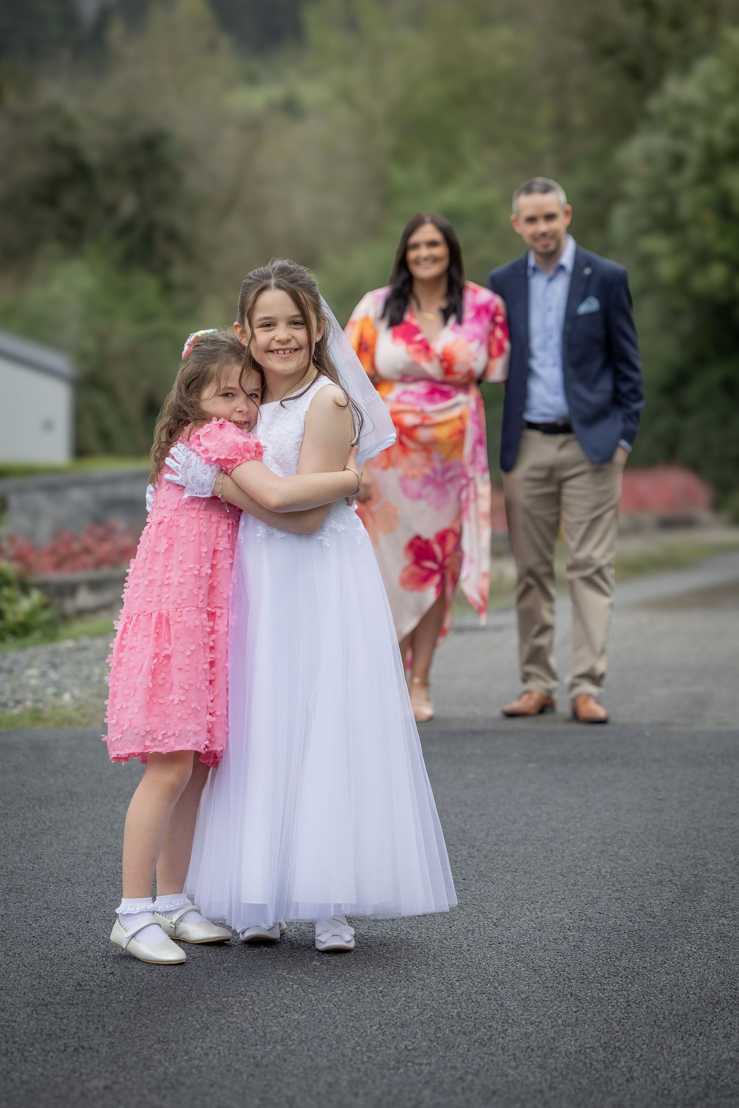 Two girls in formal dresses hug outdoors while parents stand behind them on a path.