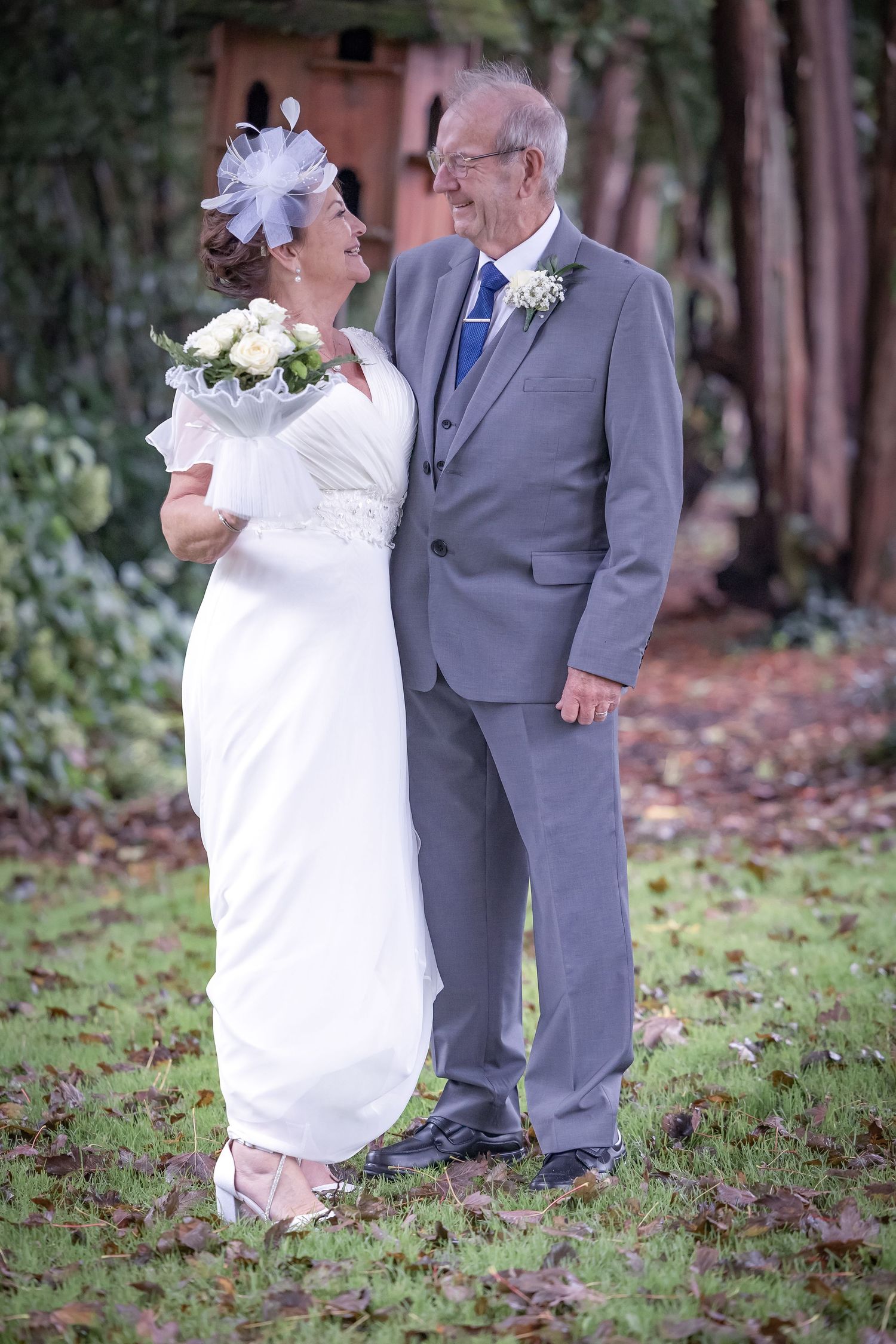 A couple in wedding attire walking together on fallen leaves in an outdoor garden setting.