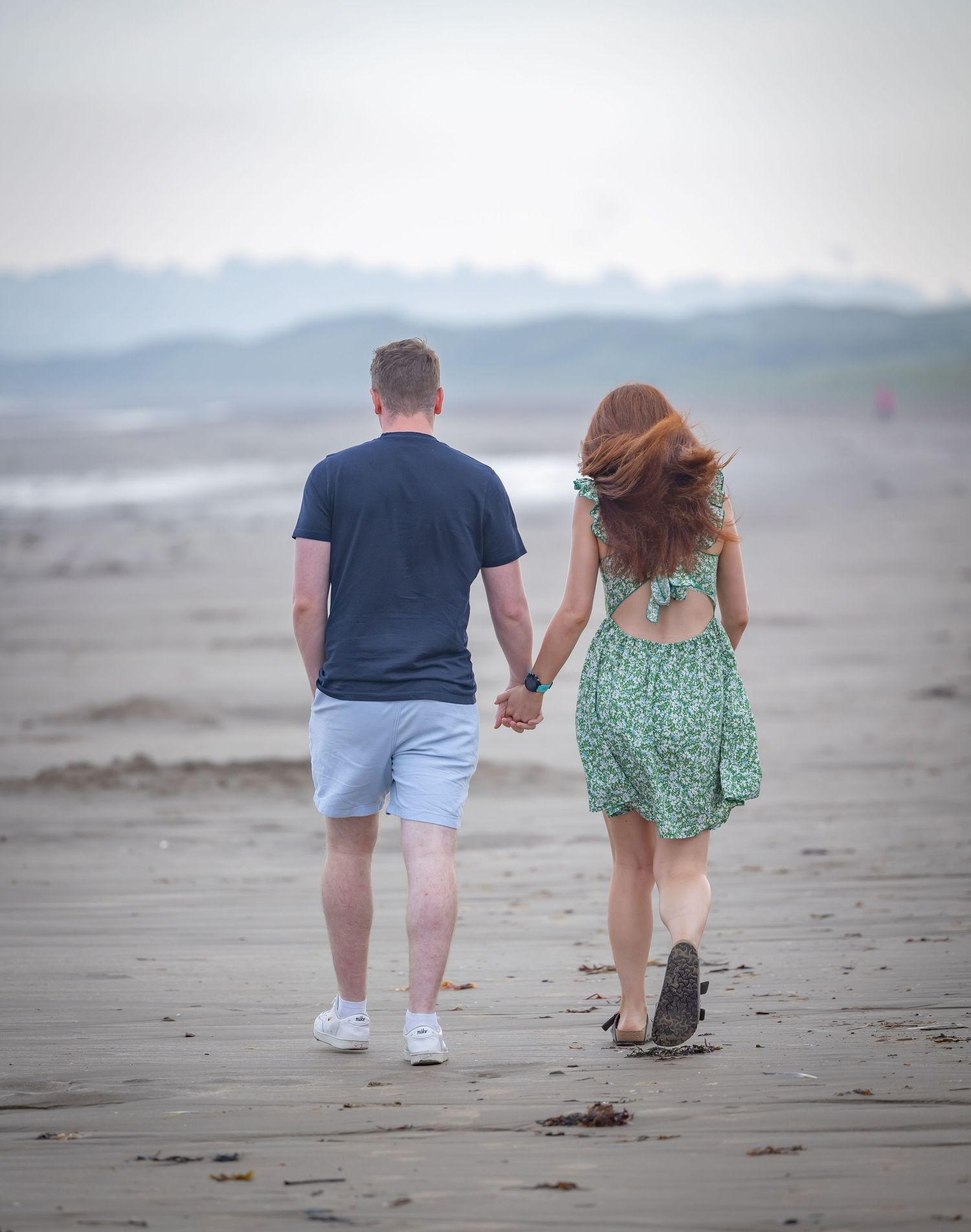Couple holding hands walking on a misty beach, woman in green floral dress with red hair, man in navy shirt and shorts.