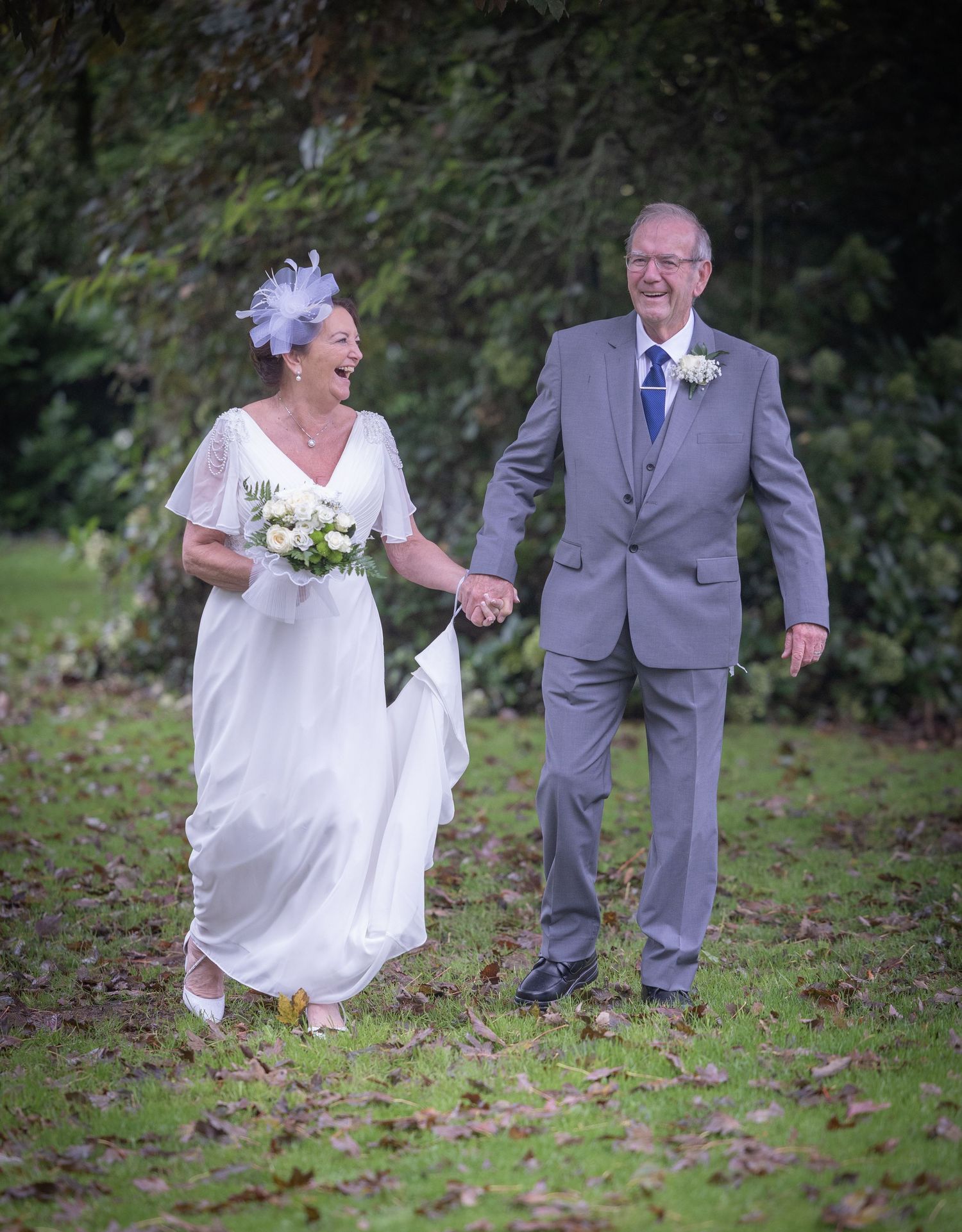 A couple in wedding attire walking and laughing together on grass with trees in the background.