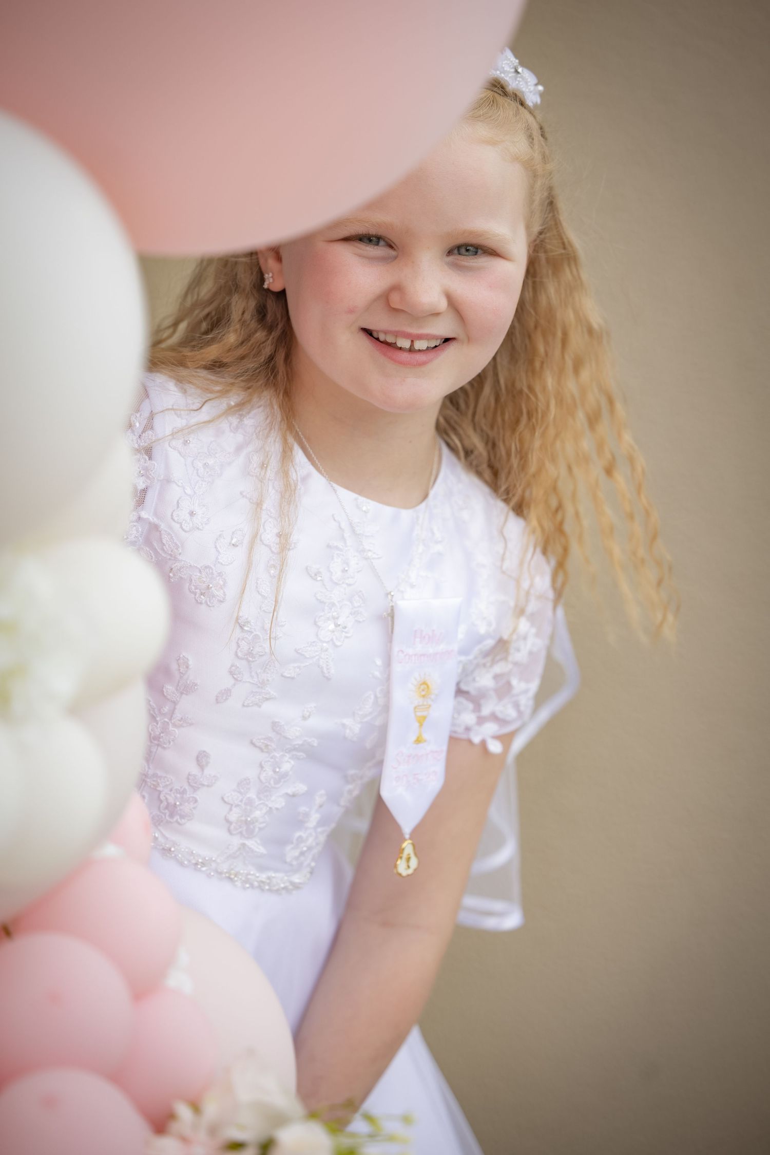 Young child in white lace dress with curly hair and pink balloons in soft focus portrait.