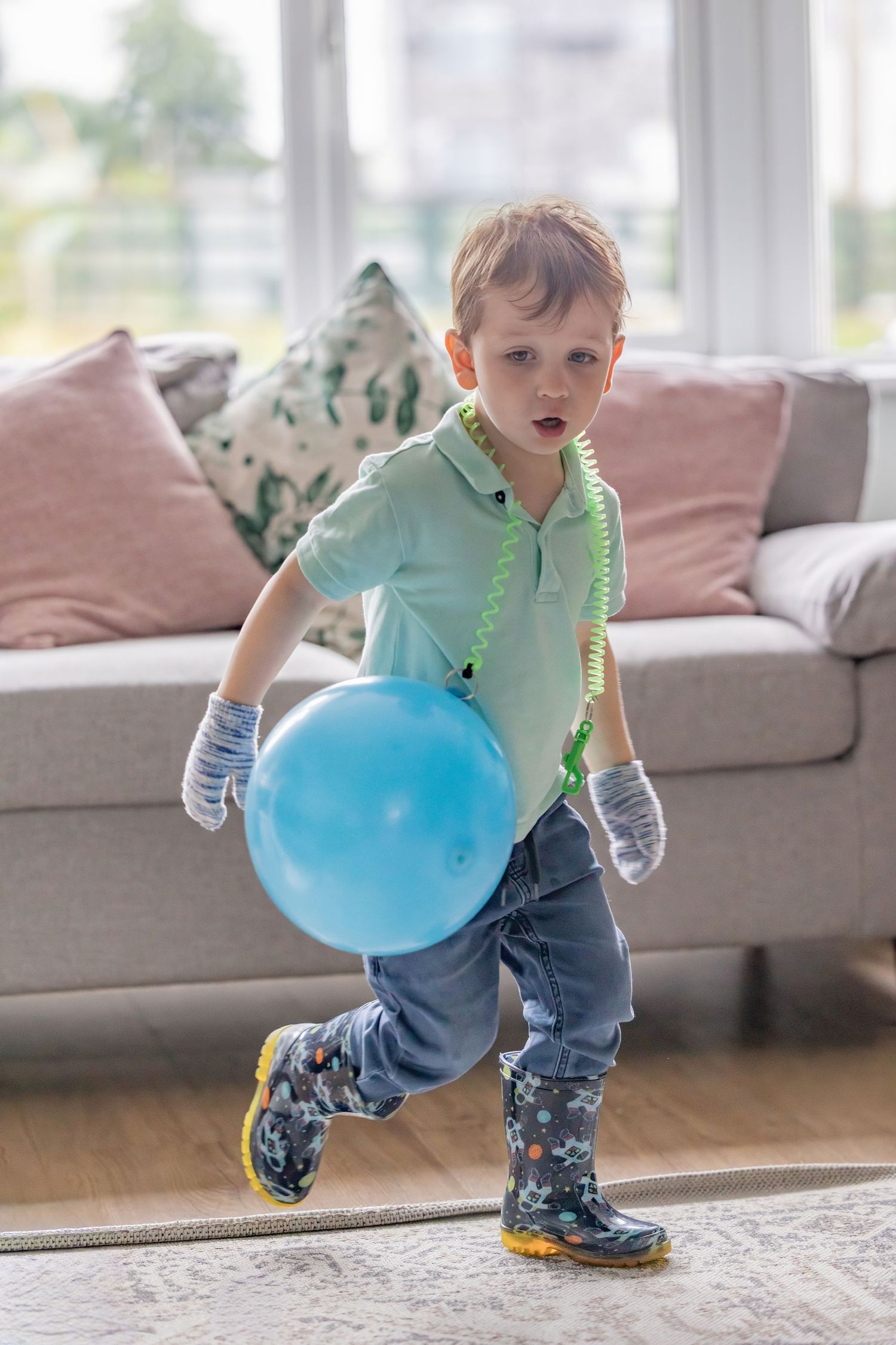 A toddler in blue shoes plays with a large blue balloon while running across a living room floor.