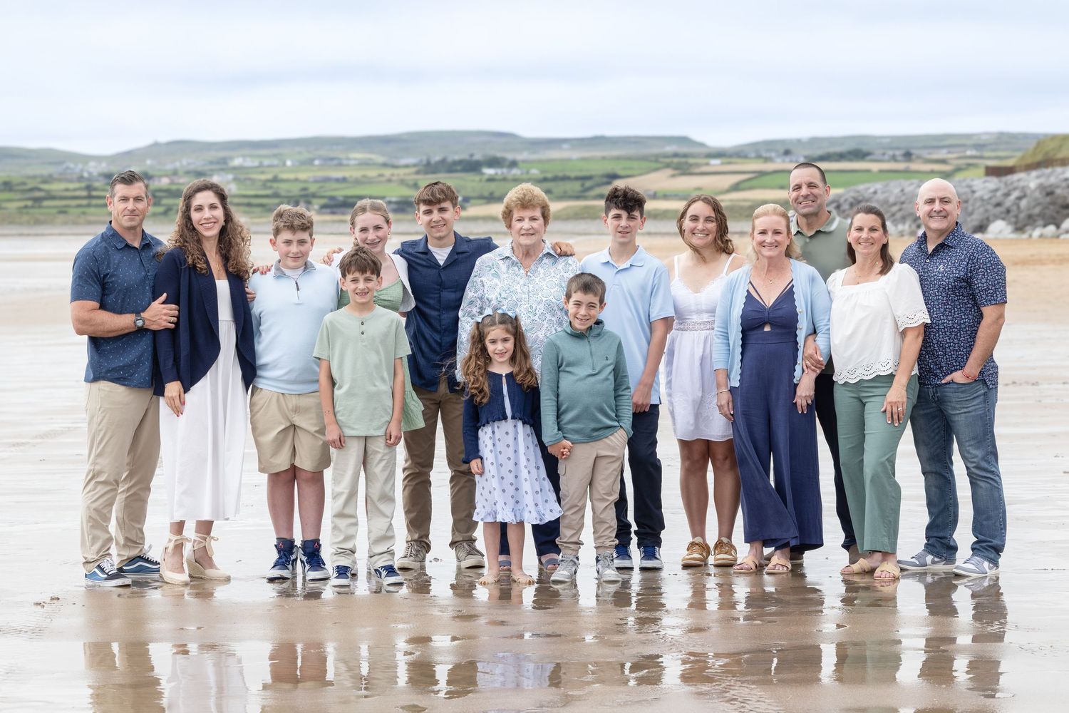 Large family group portrait on a sandy beach with green hills in the background on a cloudy day.