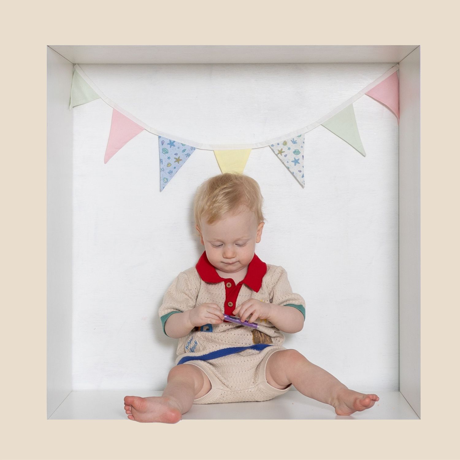 Toddler boy in beige outfit sits under colorful bunting flags on white background in framed photo.