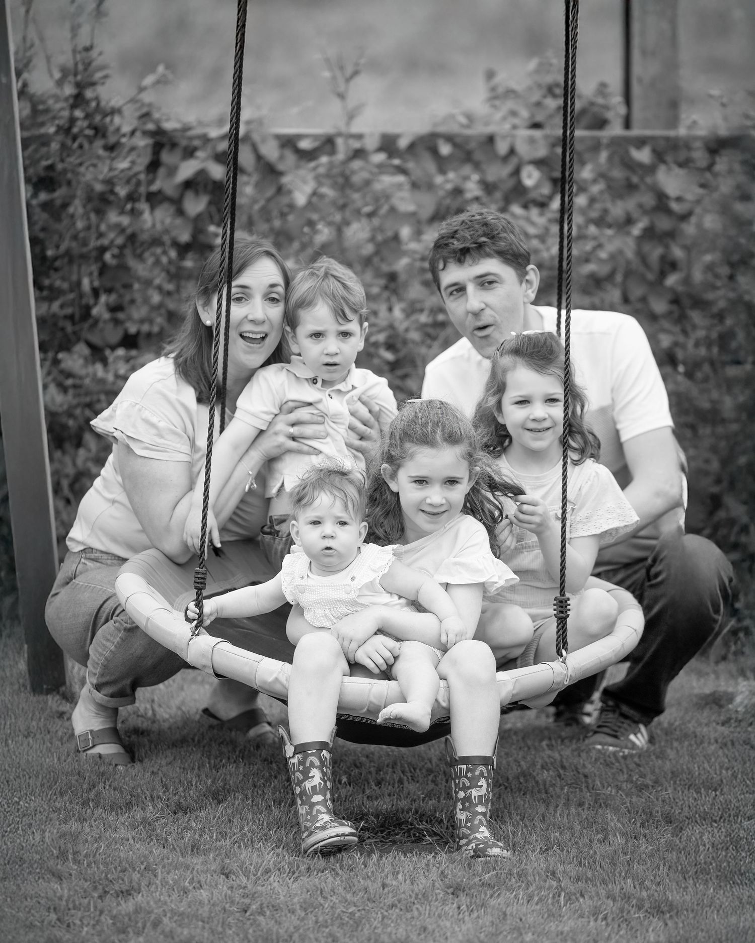 Black and white family portrait of parents with four young children gathered around a garden swing.