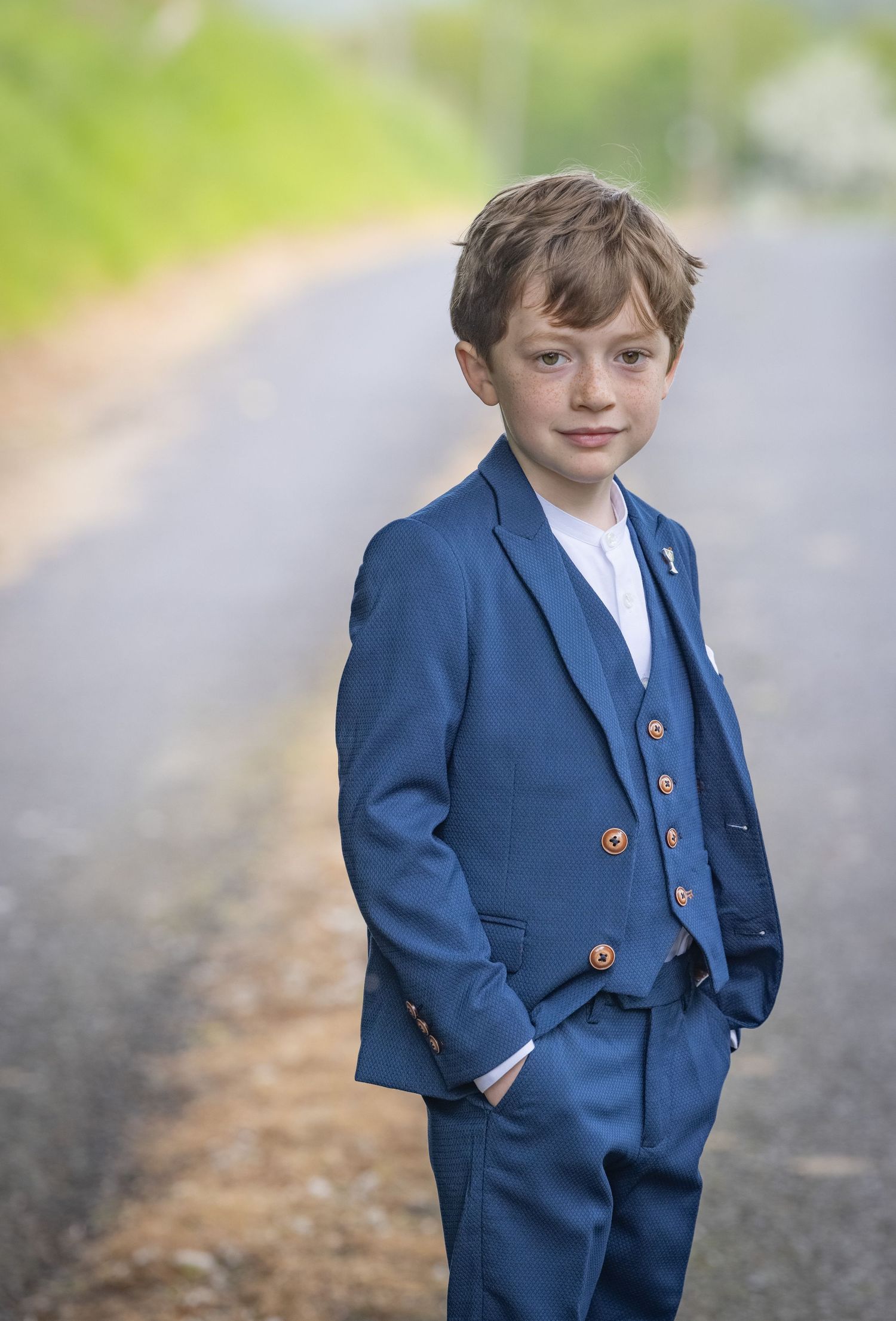 Young child wearing a navy blue suit poses playfully in an outdoor setting.