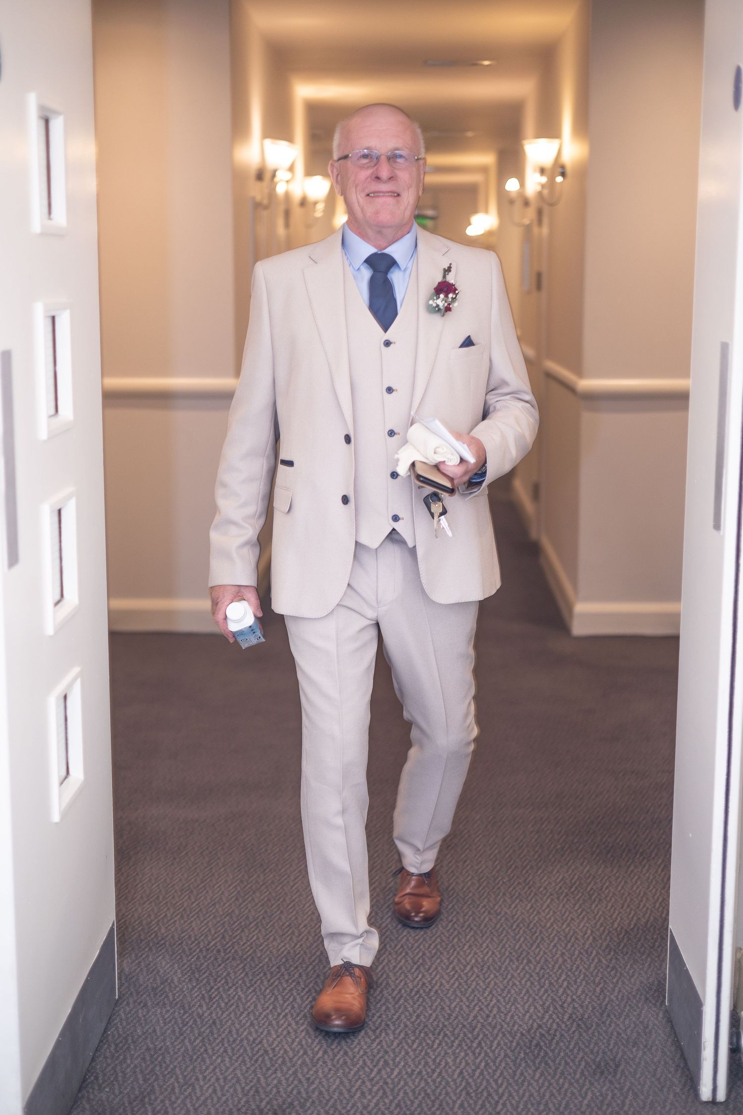 Person in light beige three-piece suit walks down hotel hallway carrying items.