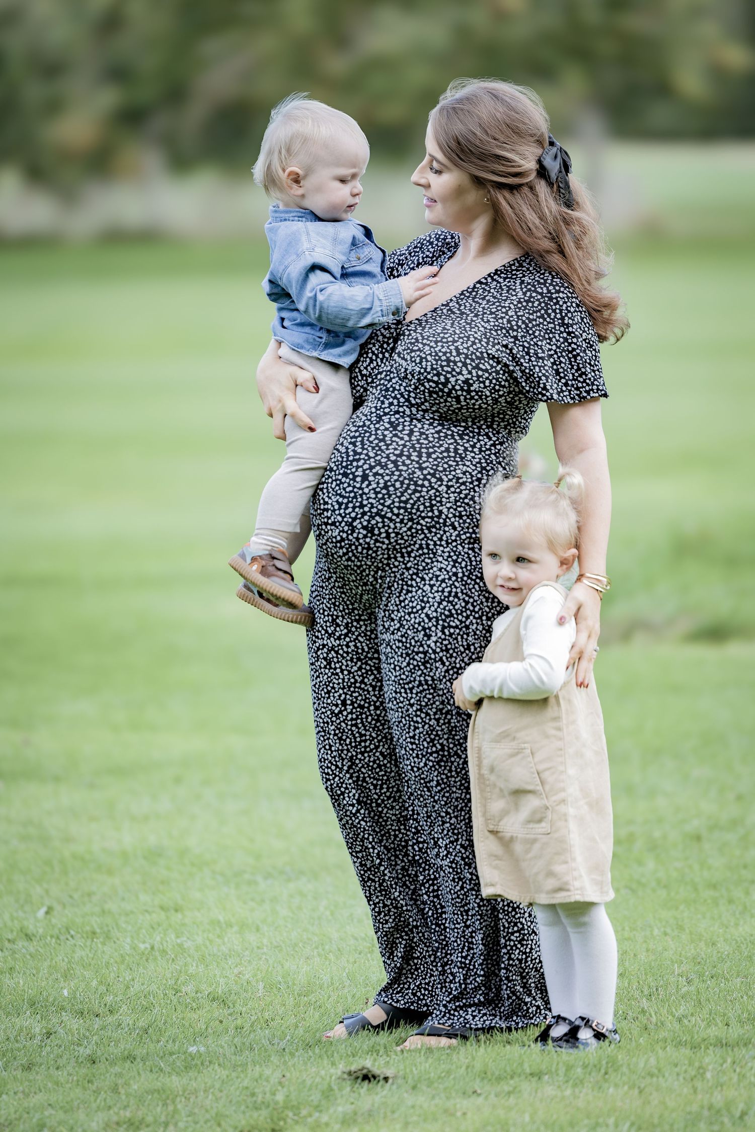 A woman in a polka dot dress holds two small children while standing in a grassy field on a sunny day.