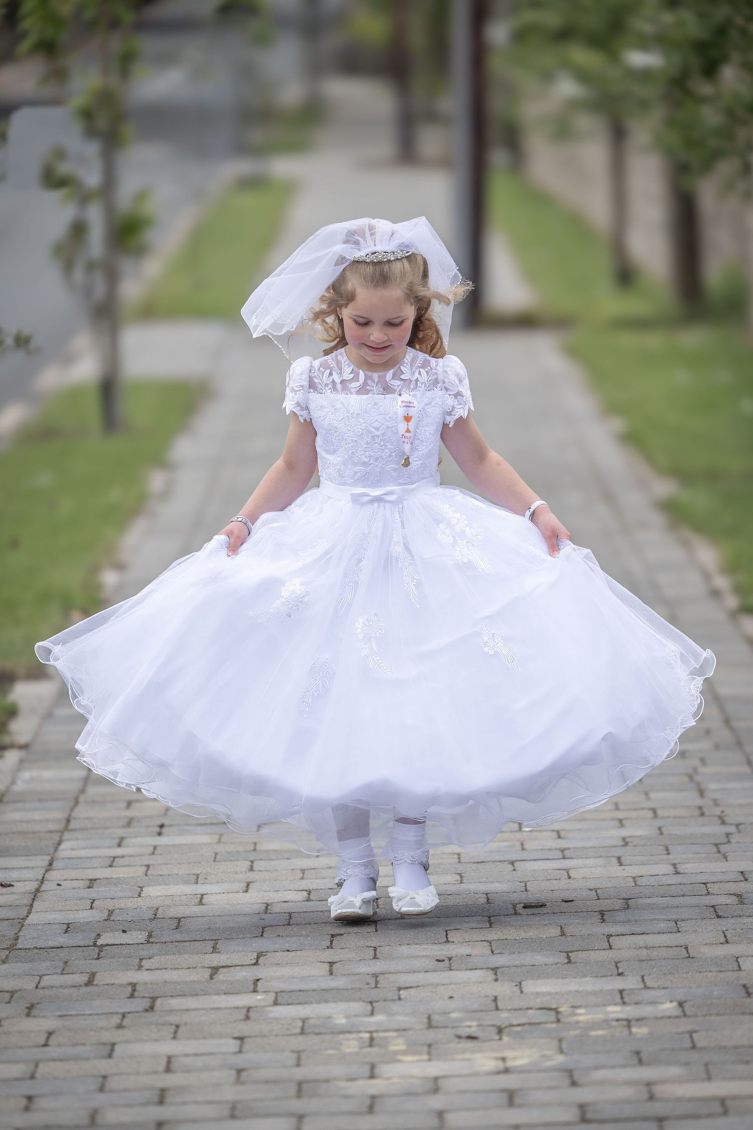 Young girl in white First Communion dress and veil twirling on a brick path outdoors.