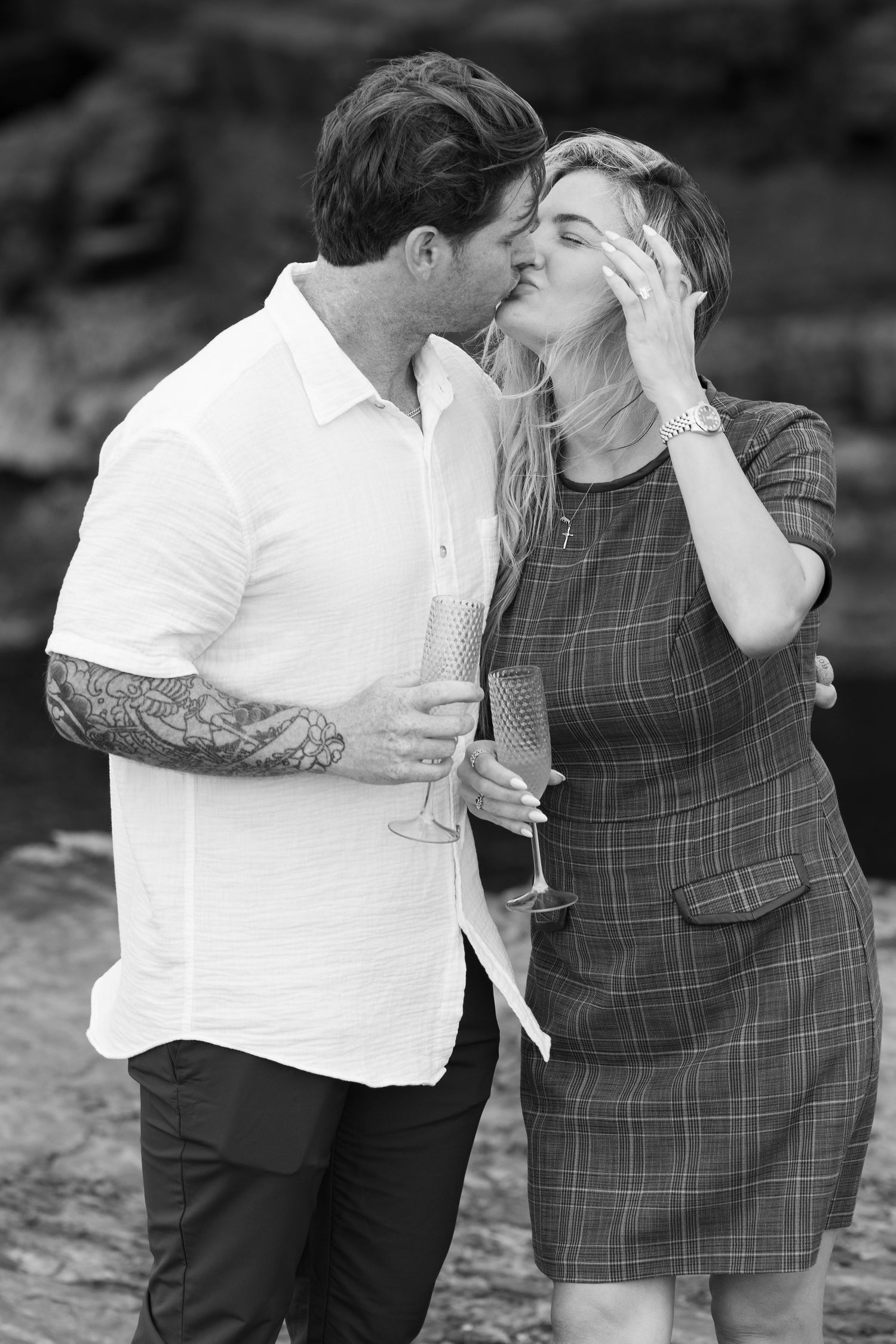 Black and white photo of a couple kissing while holding champagne glasses outdoors by rocky terrain.
