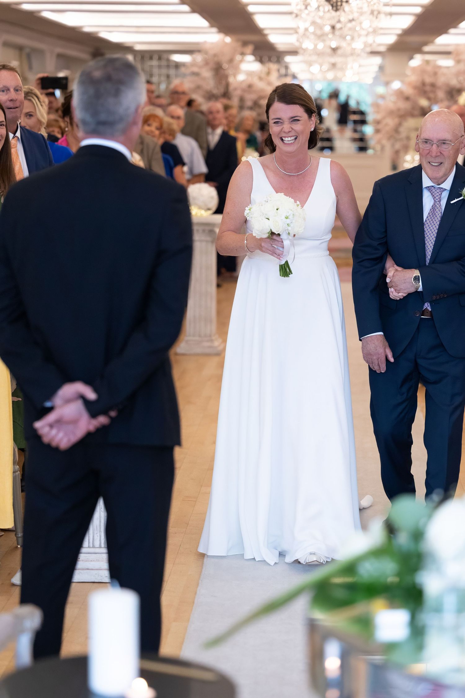 Smiling bride in white gown walks down the aisle holding white bouquet, escorted by a man in a blue suit.
