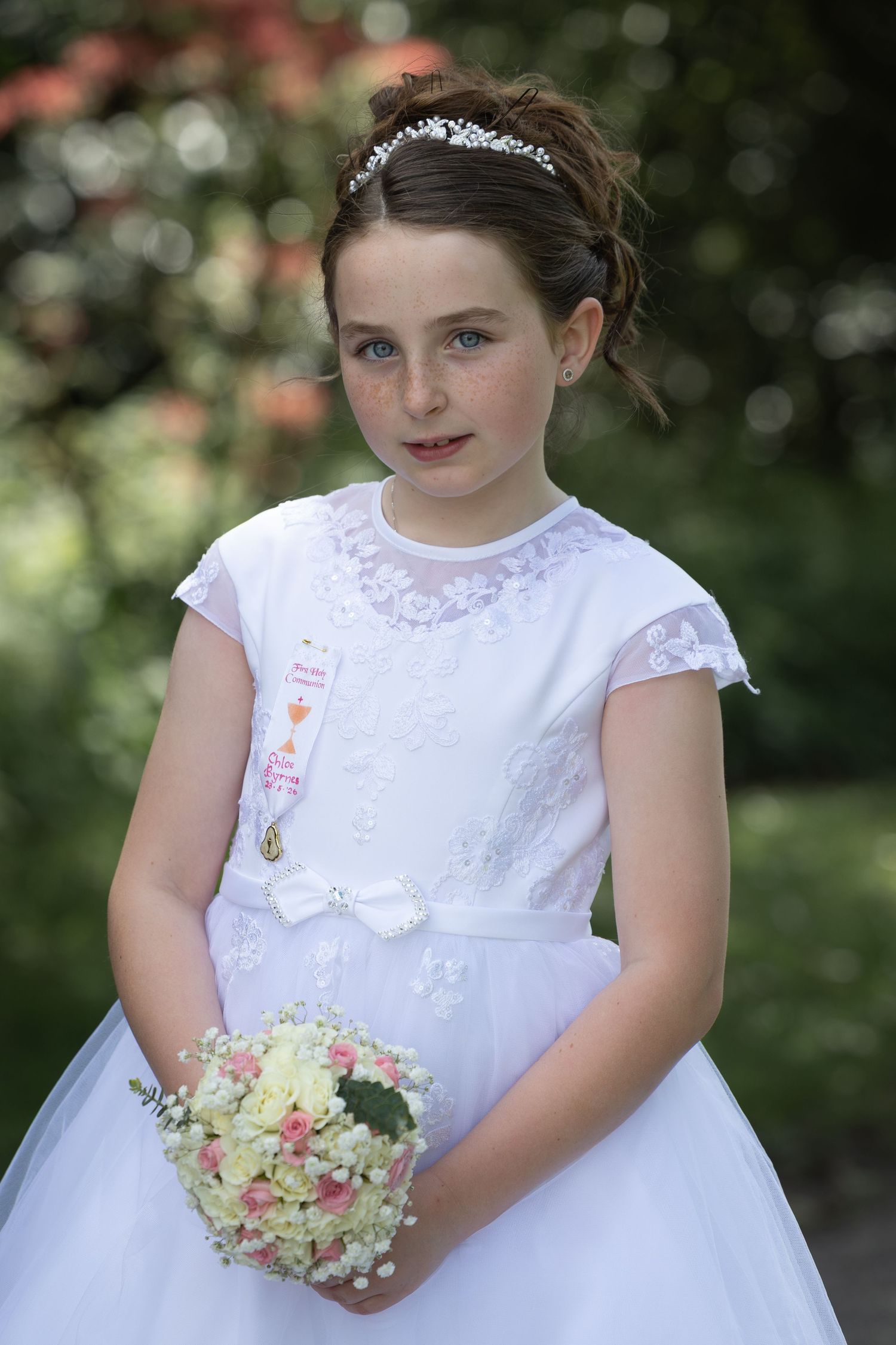 Young girl in white First Communion dress holding bouquet, wearing tiara and religious medal, posing outdoors.