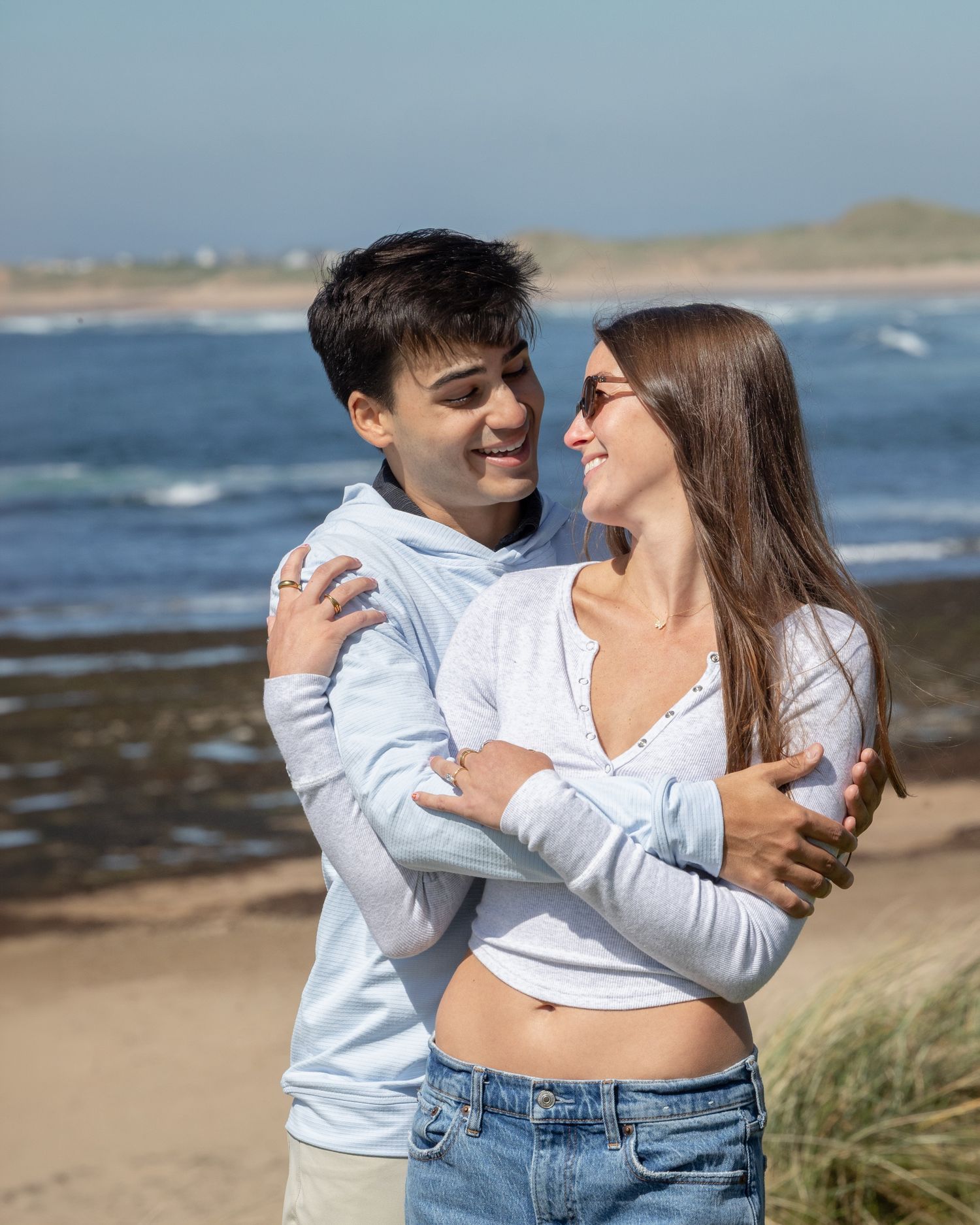 Happy young couple embracing and smiling on a sunny beach with ocean waves behind them.