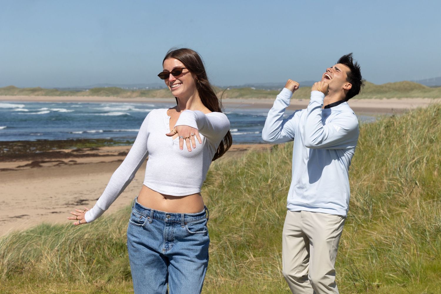 Young couple enjoying a sunny beach day, woman in white crop top and jeans, man in light blue shirt laughing outdoors.