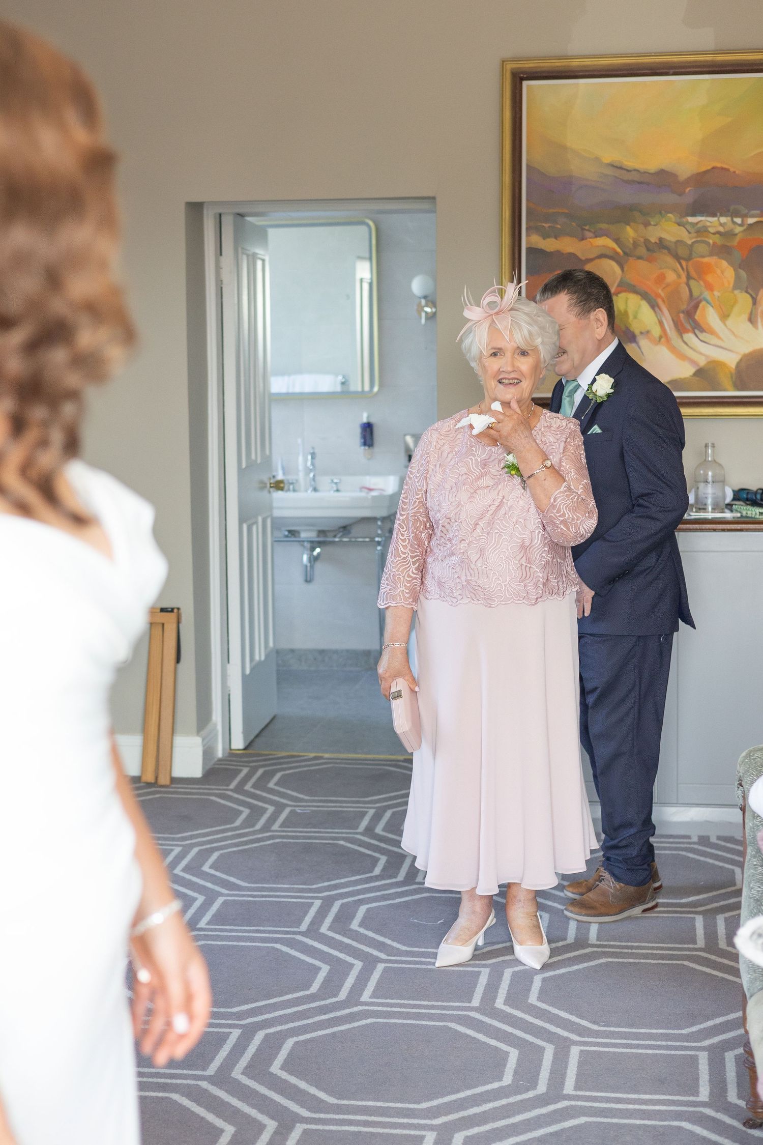 Woman in pink lace dress and man in suit stand in room with geometric patterned carpet and landscape painting.