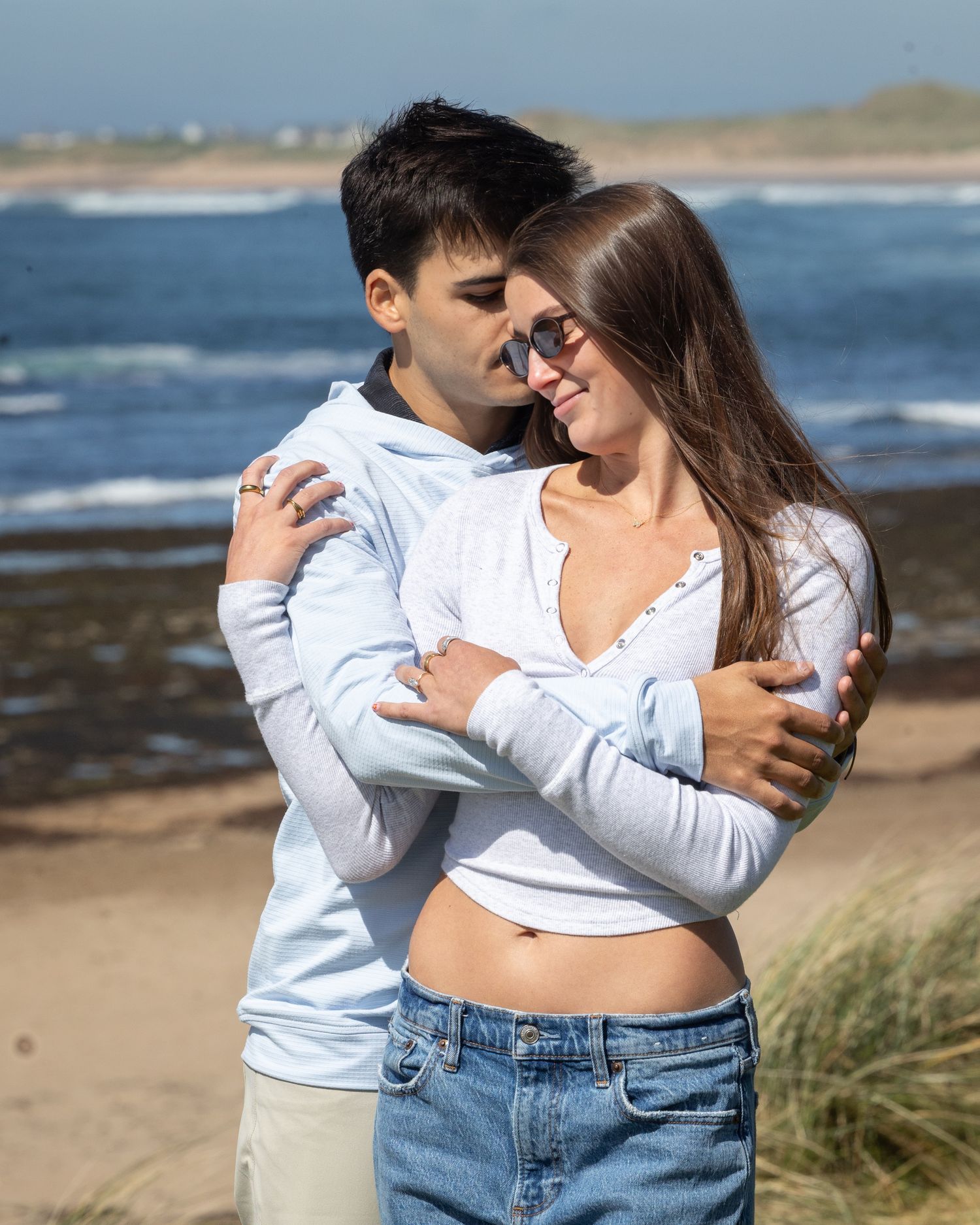 A young couple embracing on a sunny beach, wearing white hoodies and jeans, smiling warmly with ocean waves in the background.