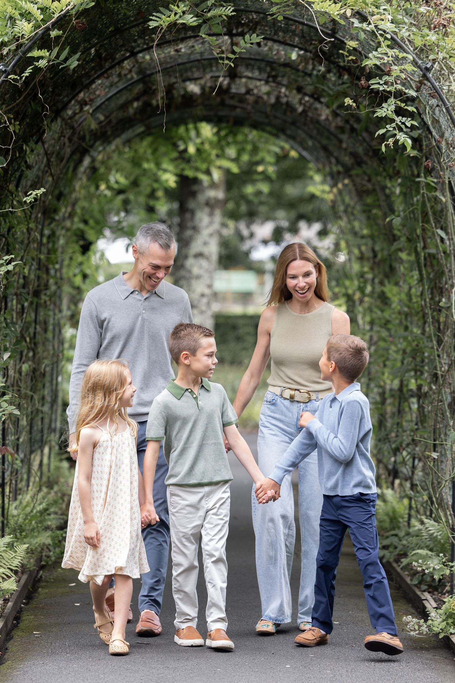 Happy family of five walking under a lush green garden archway, smiling and holding hands outdoors.