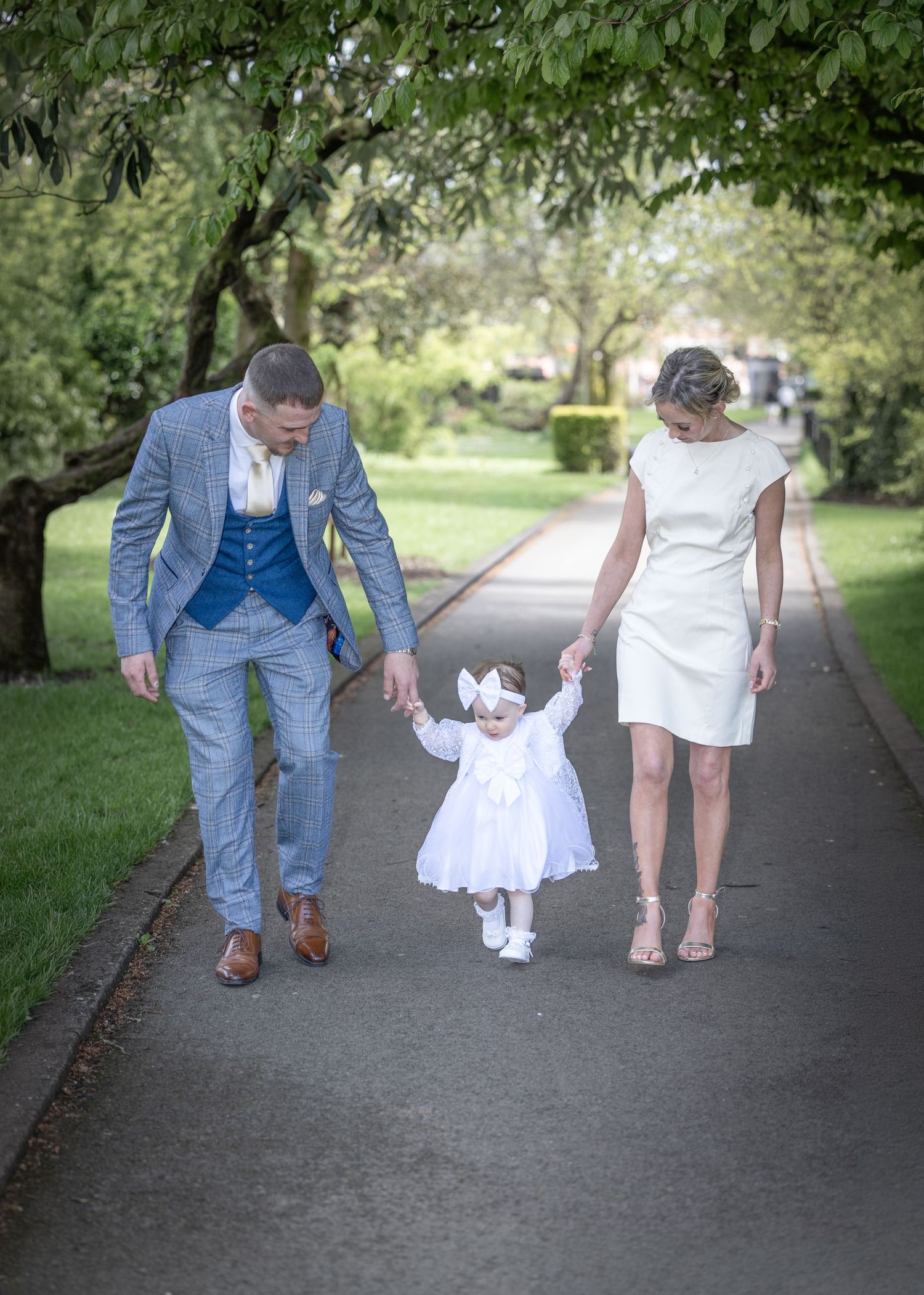 A couple in formal attire walks hand-in-hand with their toddler in a white dress along a tree-lined path.