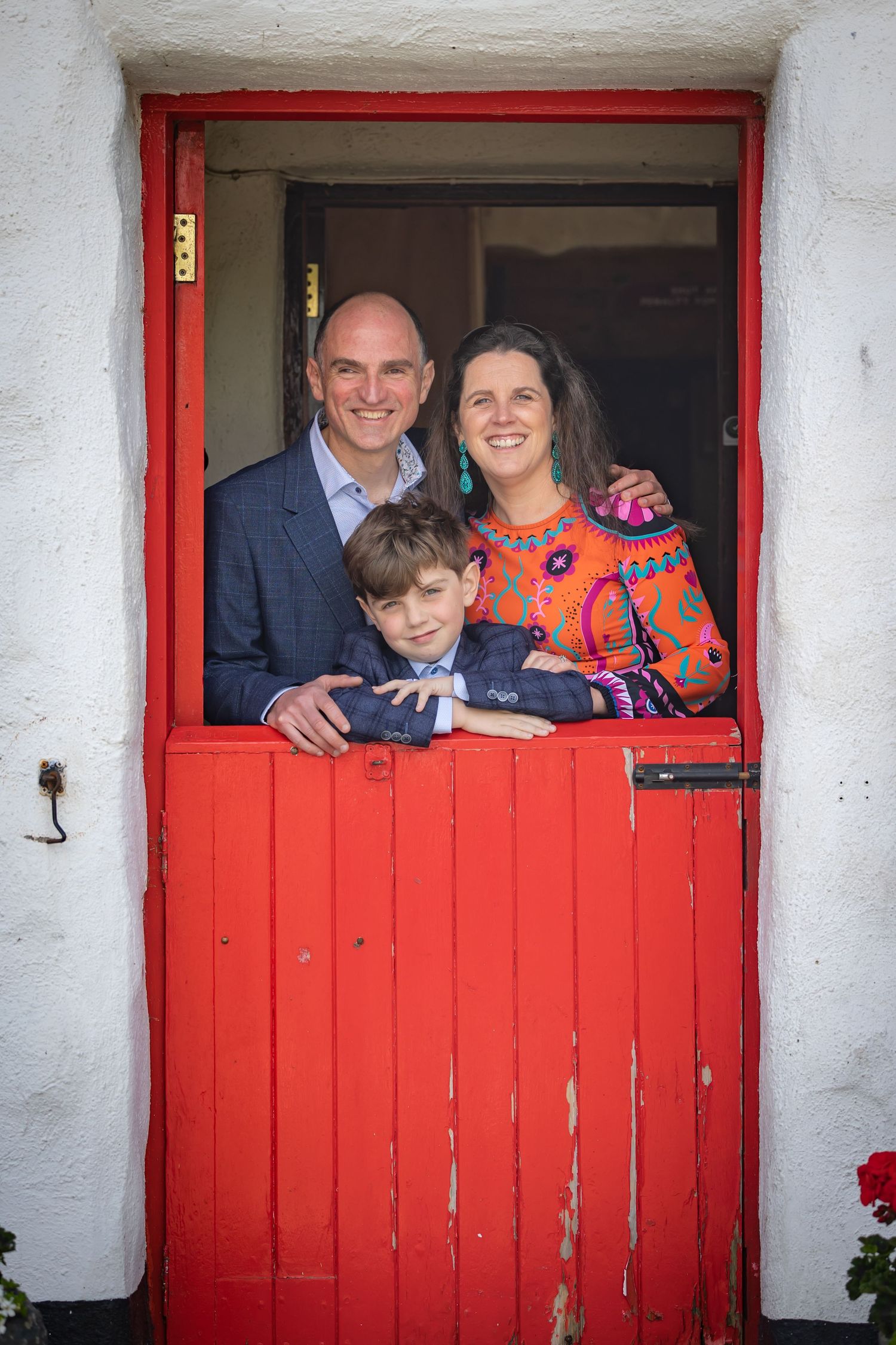 Family members peek through a bright red stable door in a white wall, creating a playful and charming portrait.