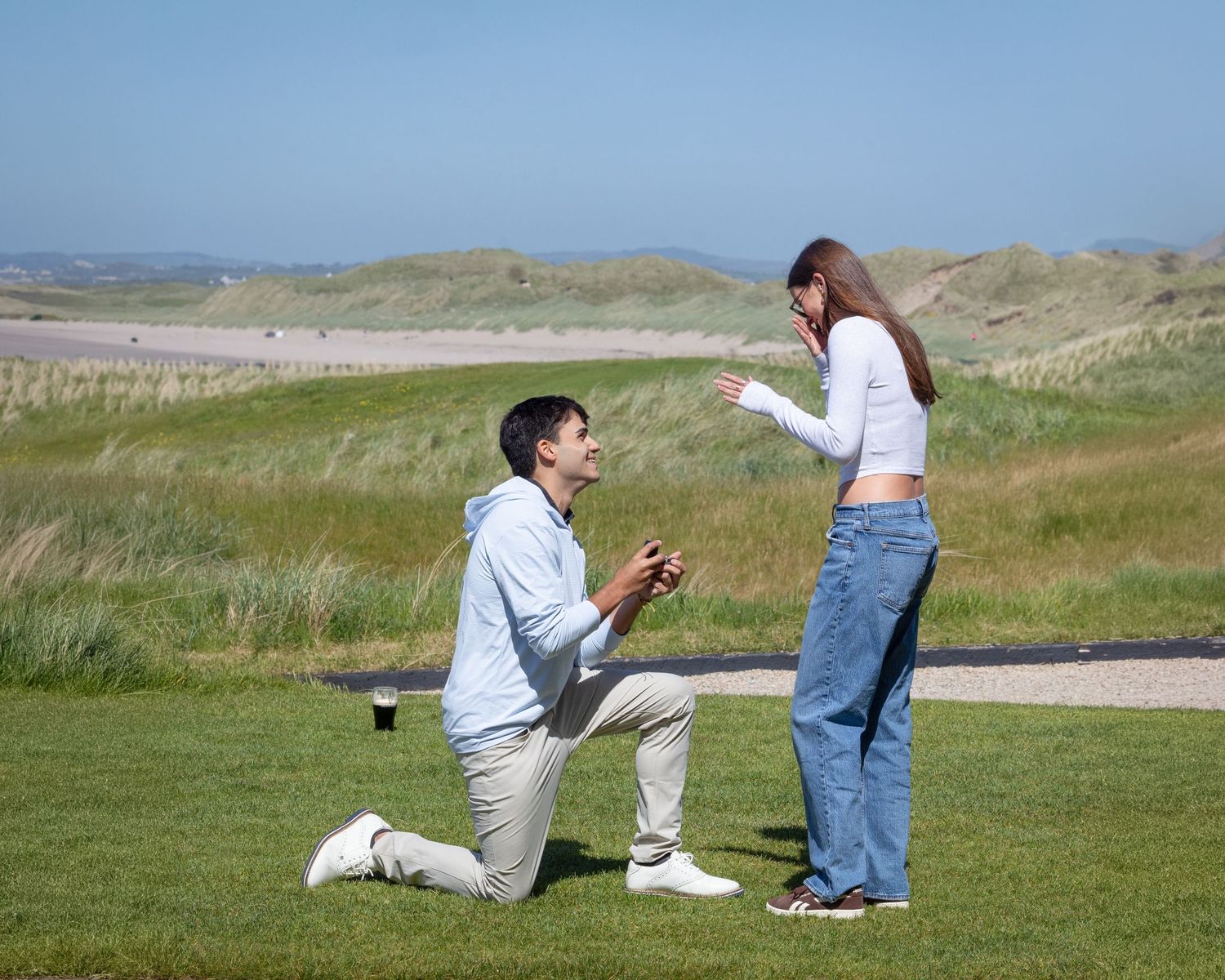 Man proposes on one knee to woman outdoors on green grass with sandy dunes and blue sky in background.