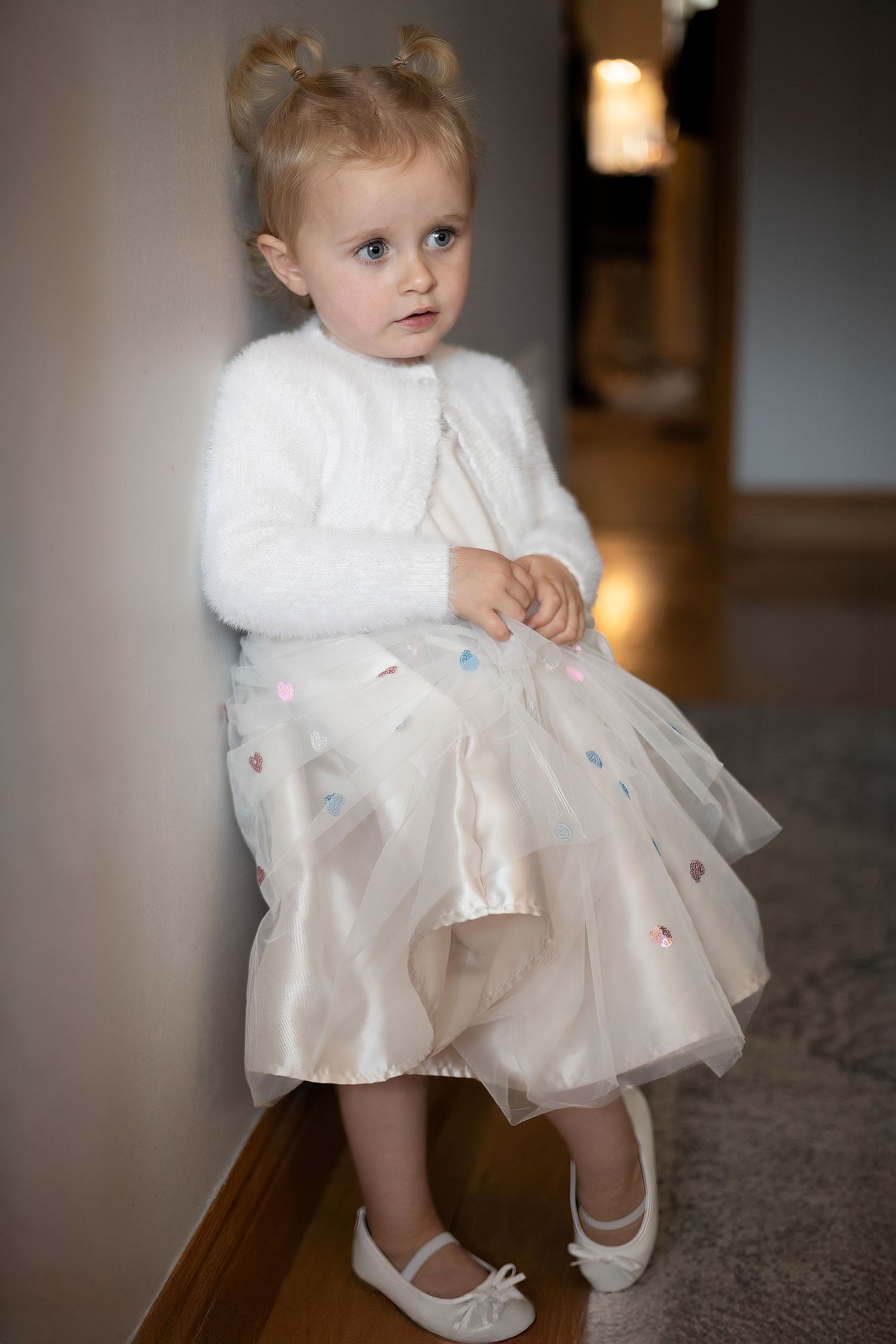 A small figure in a white tulle dress and shoes sits on dark flooring in a dimly lit interior space.