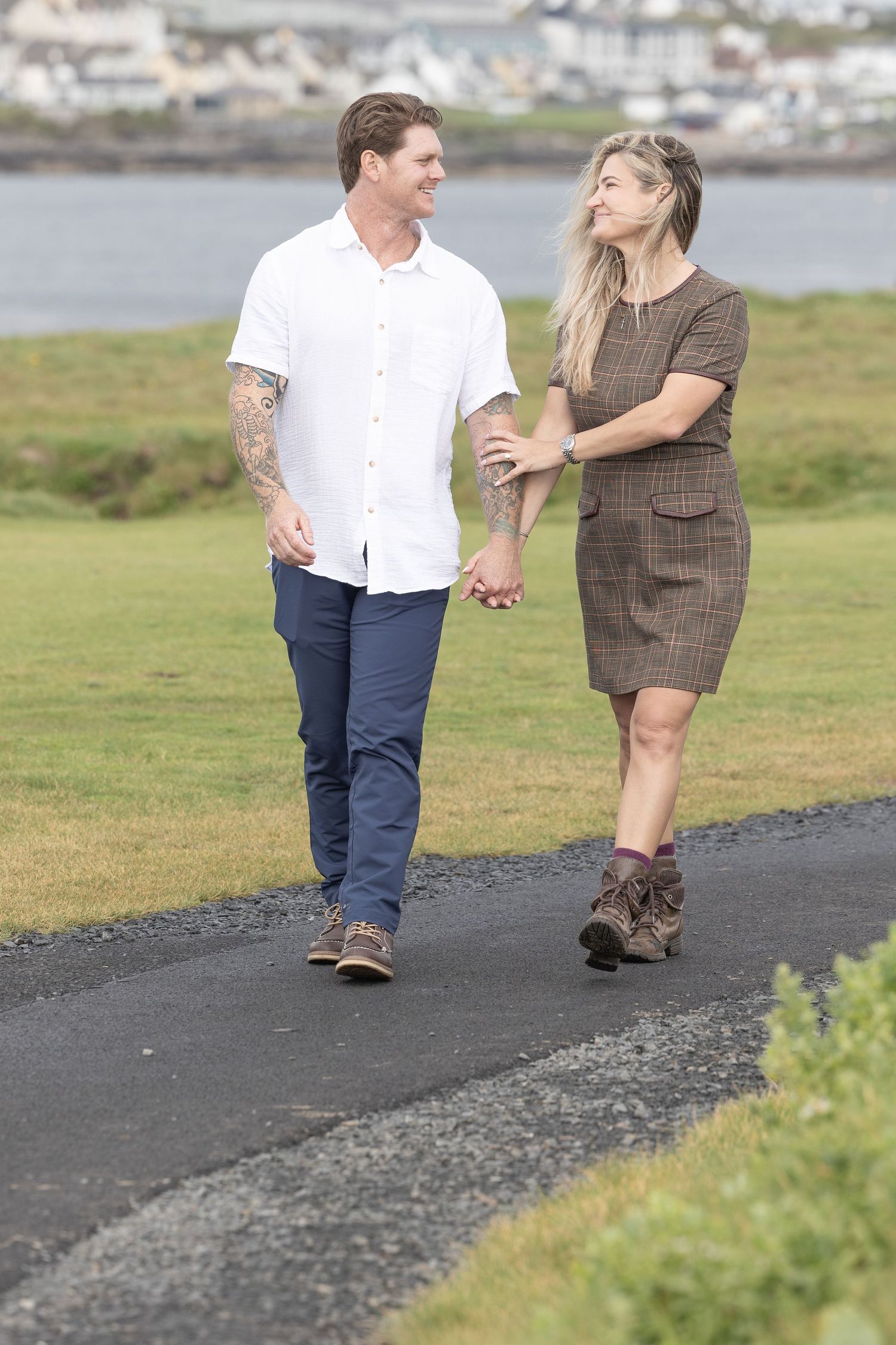 Two people walking together on a paved coastal path with water and grassy areas visible in the background.
