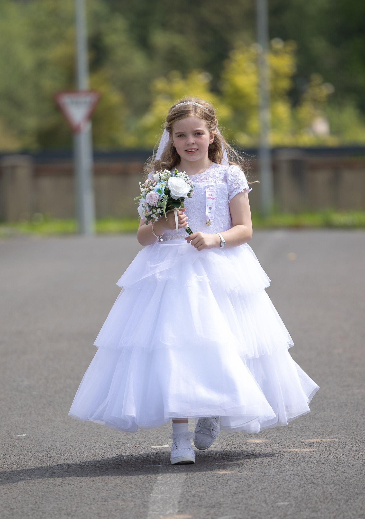Child in white formal dress sits gracefully on pavement with floral accessories.