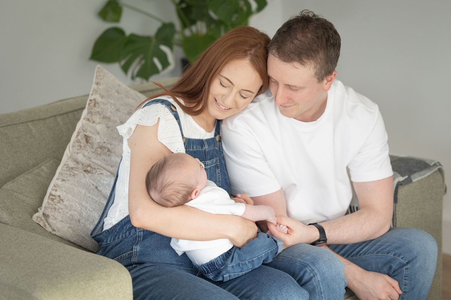 A loving family sits together on a couch, sharing tender moments with their young baby in a bright room.