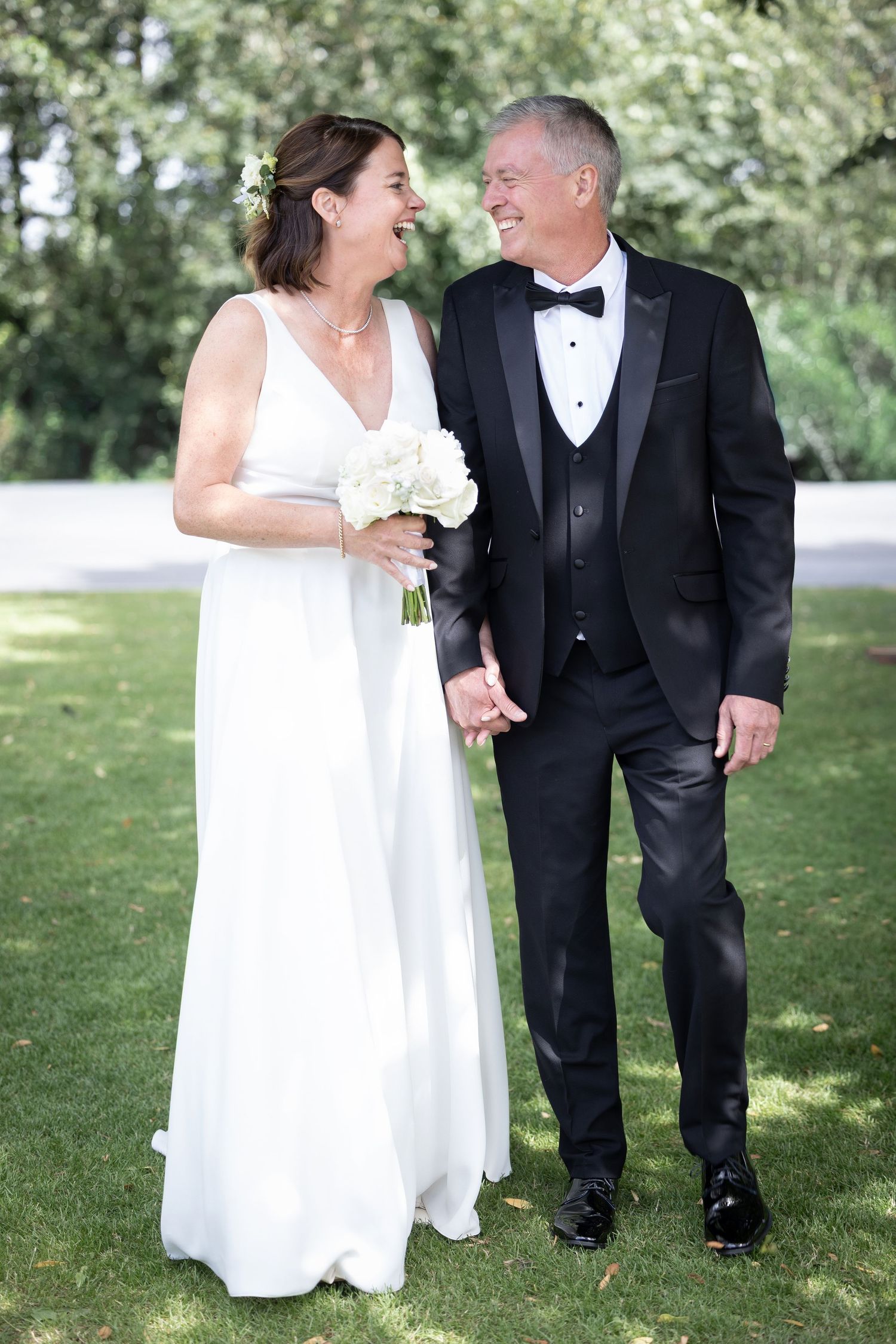 Happy mature bride and groom holding hands, walking outdoors on grass, smiling at each other on their wedding day.