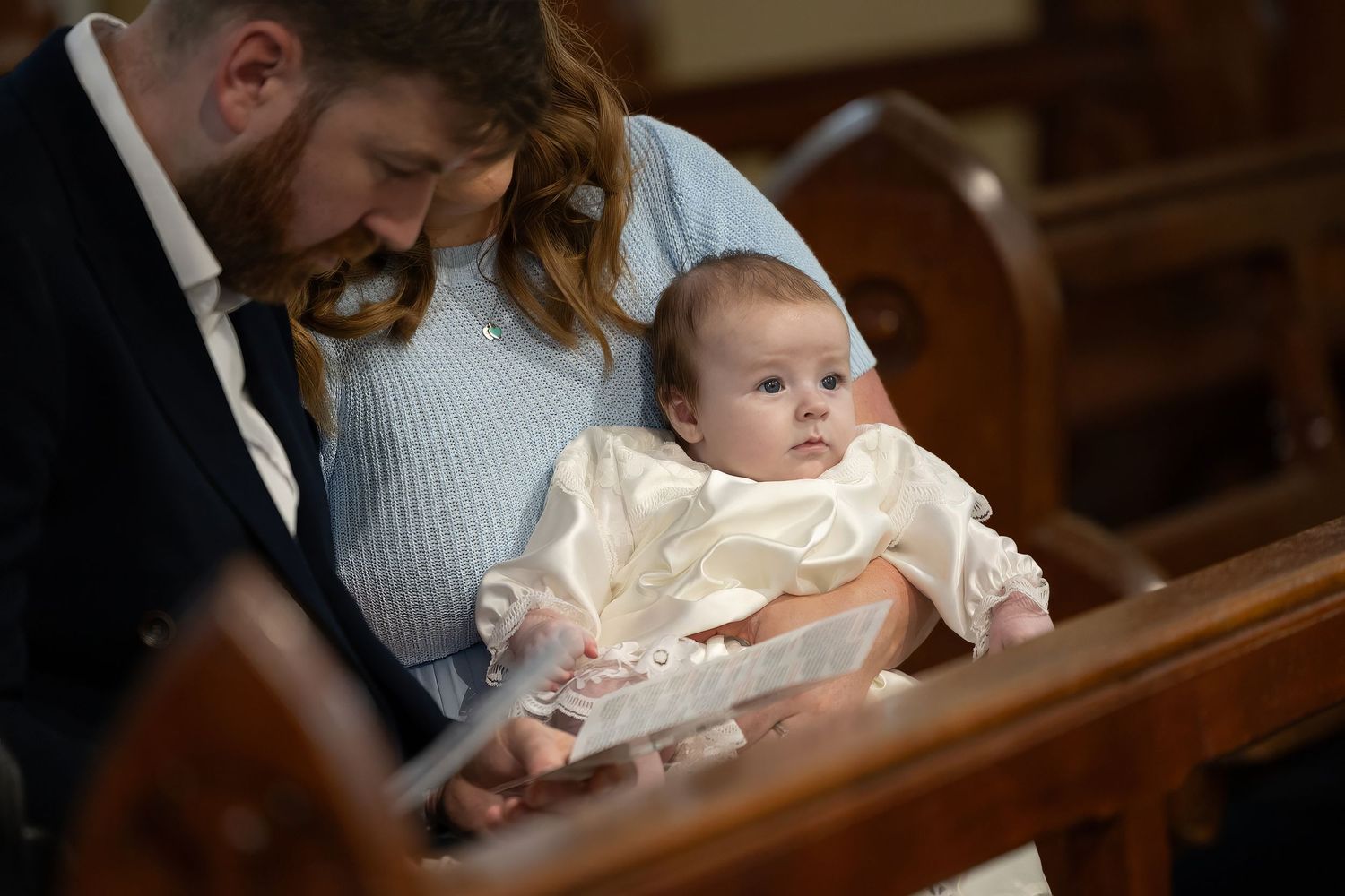 Parents holding a baby in a white christening gown during a baptism ceremony in a church pew.
