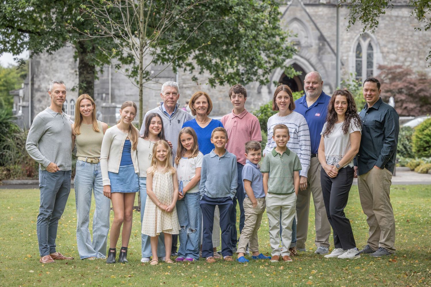 Large multigenerational family group portrait outdoors on green lawn with historic stone church in background.