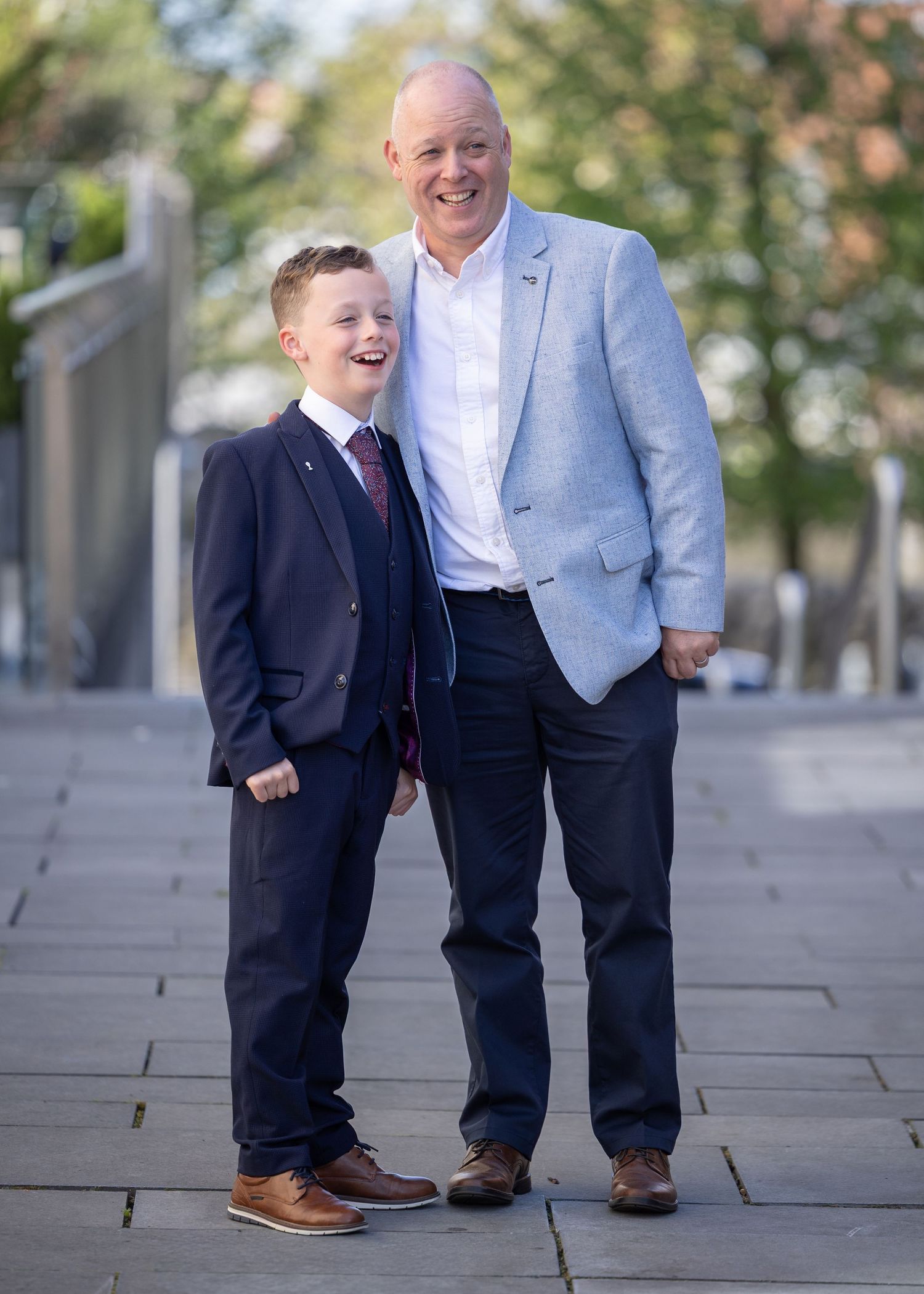 A smiling boy in a navy suit and man in light blue blazer pose together on a stone pathway outdoors.