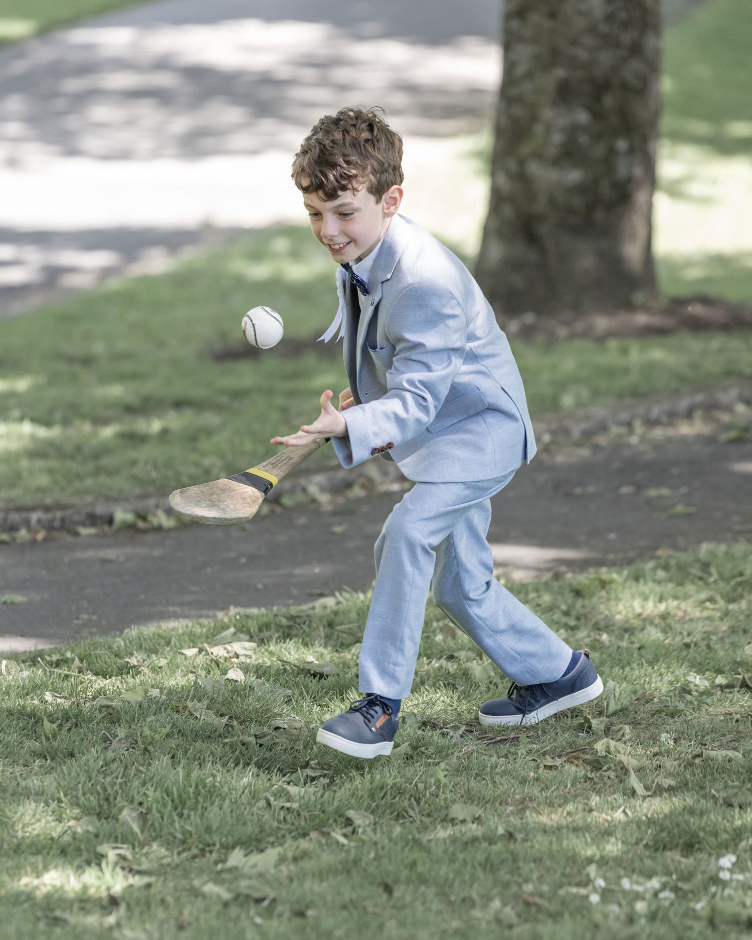 A young child in light blue clothing chases after a ball on a grassy area under dappled sunlight.