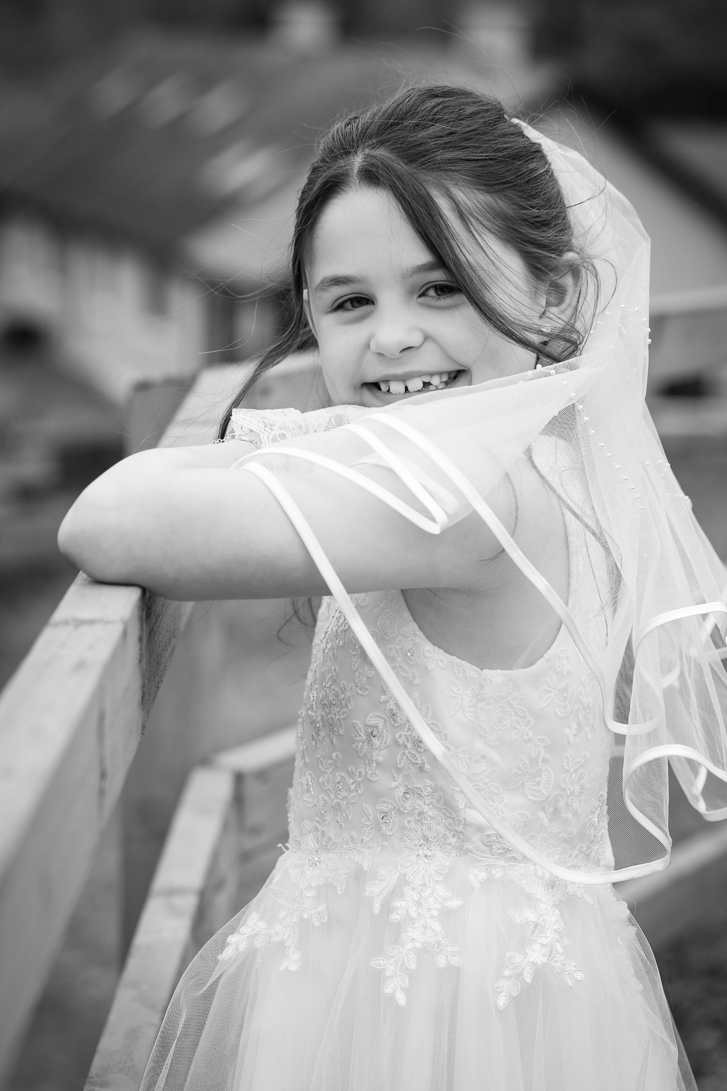 A smiling young girl in a white lace dress and veil leans on a wooden fence in a black and white photo.