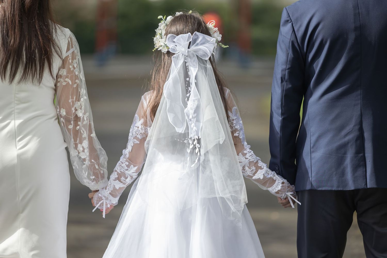 Young girl in white First Communion dress and floral veil holds hands with parents outdoors.