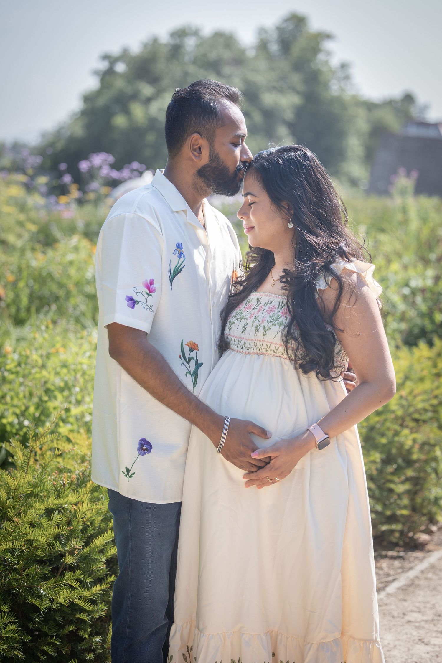 Couple in matching floral outfits sharing a tender moment outdoors, woman in white dress cradling baby bump.