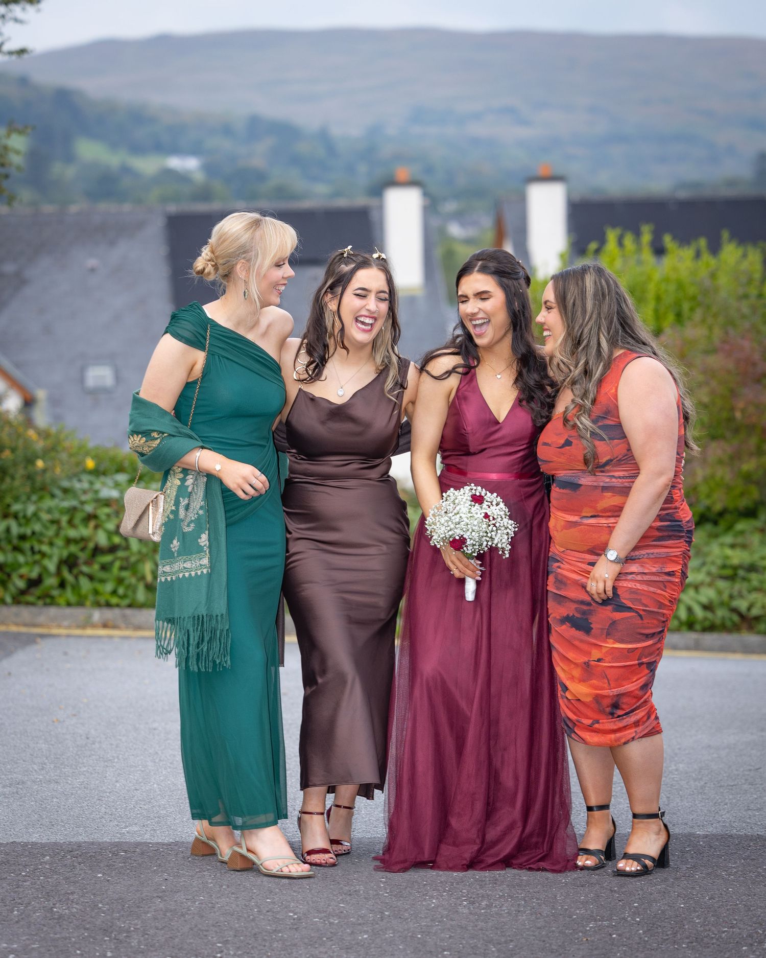 A group of friends in formal dresses laughing and sharing a candid moment outdoors with mountains in the background.