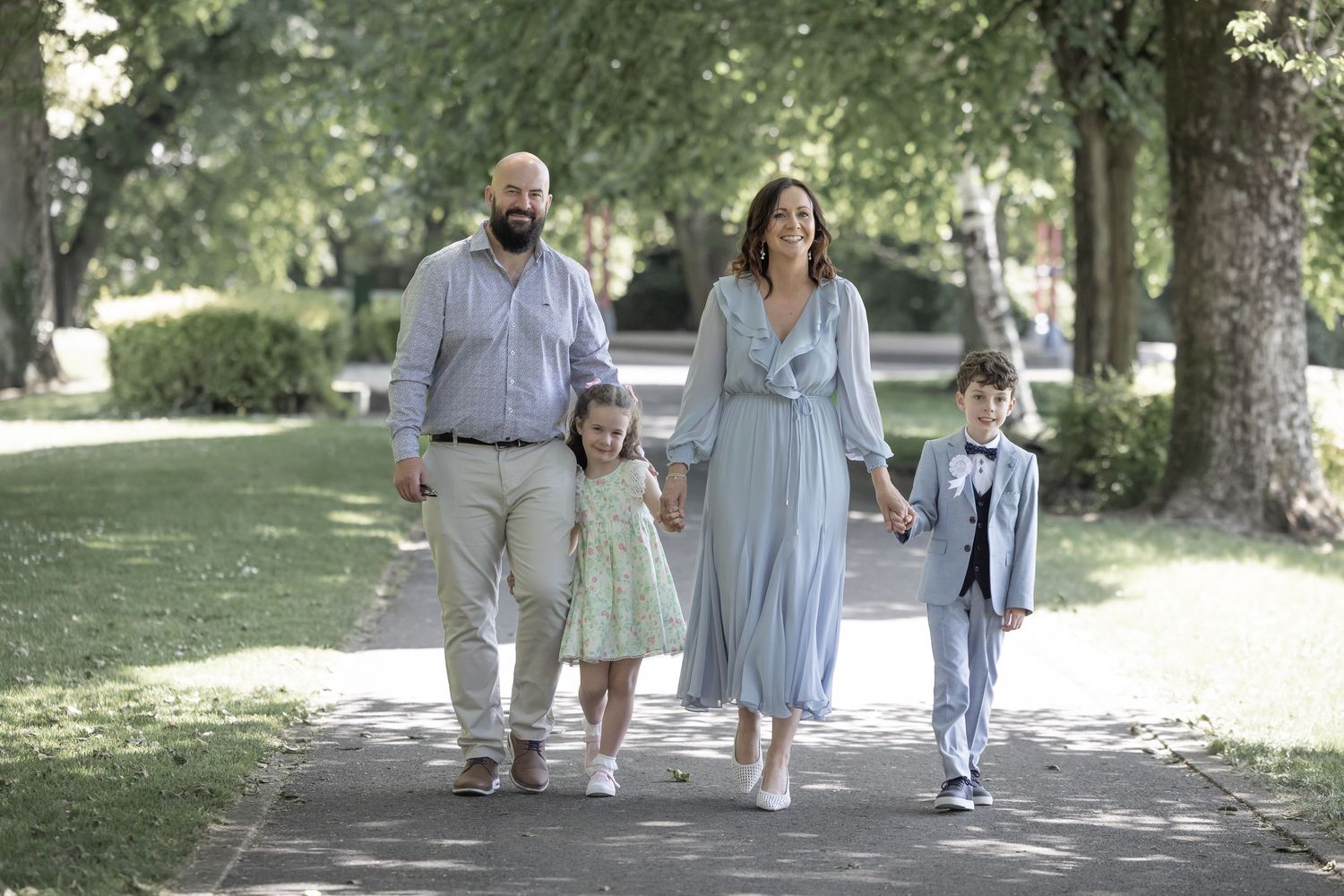 Family walking together on a tree-lined path in a park wearing coordinated light blue and grey outfits.
