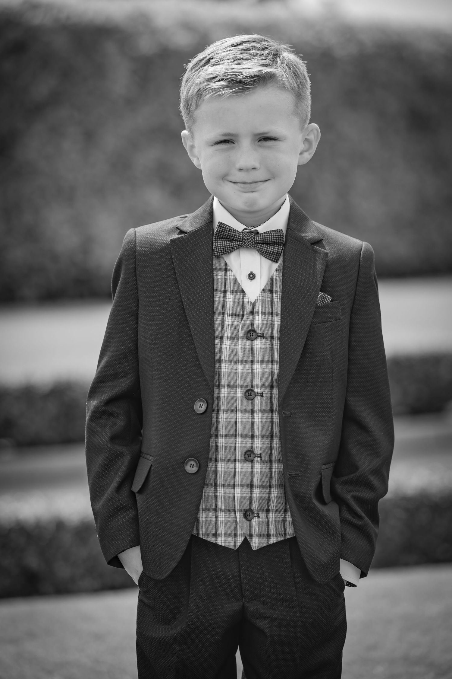 Child in formal black suit and plaid vest poses for portrait outdoors.