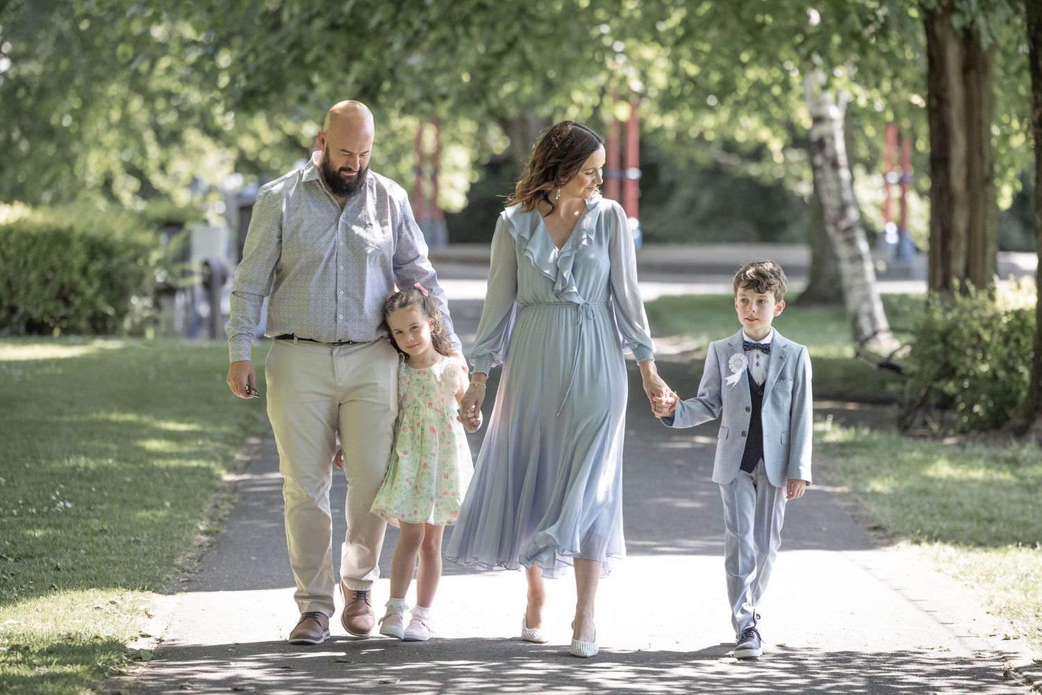 A family of four walks together on a tree-lined path in a park on a sunny summer day.