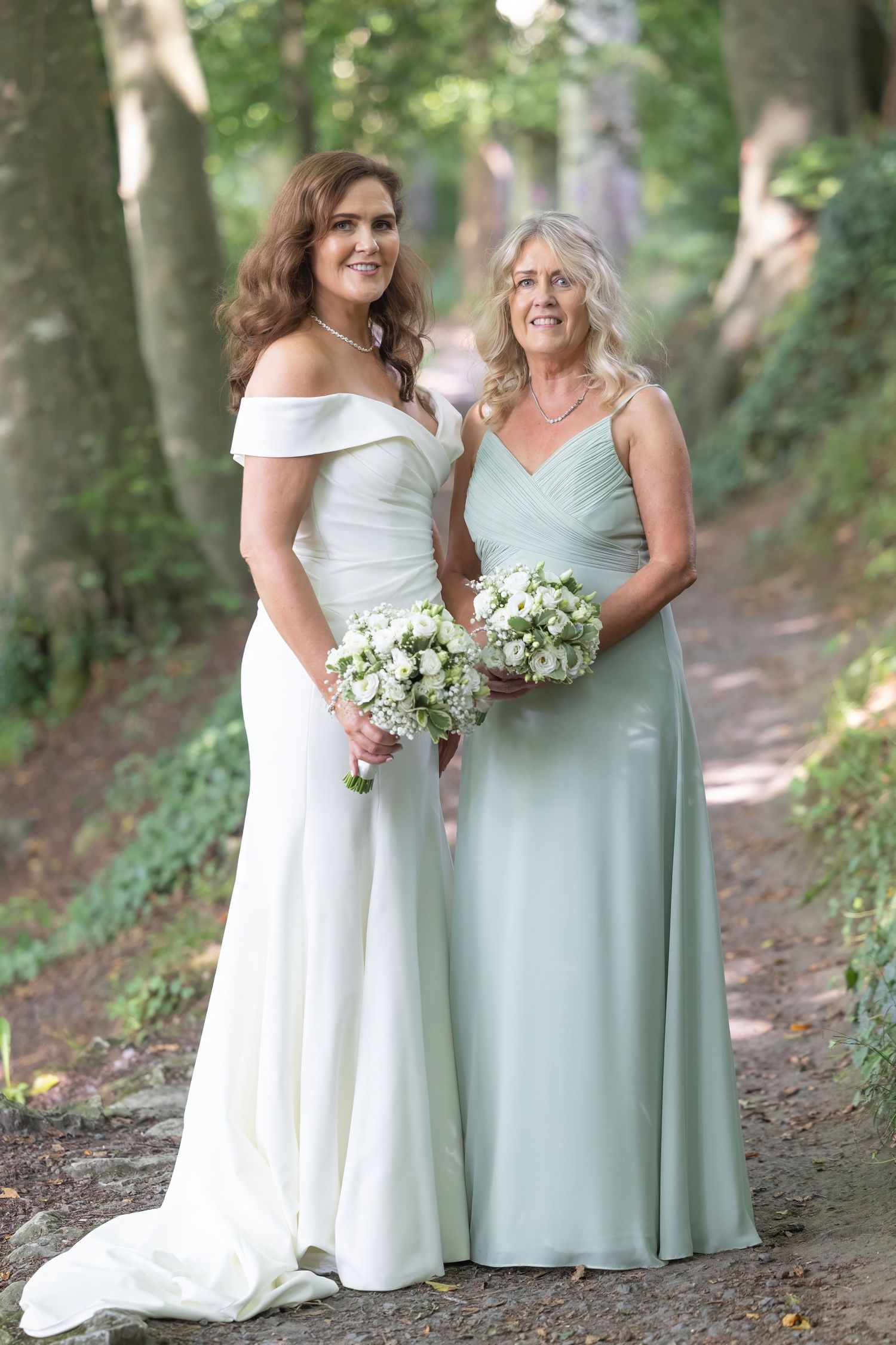 Two women in formal dresses pose outdoors holding white bouquets on a wooded path.