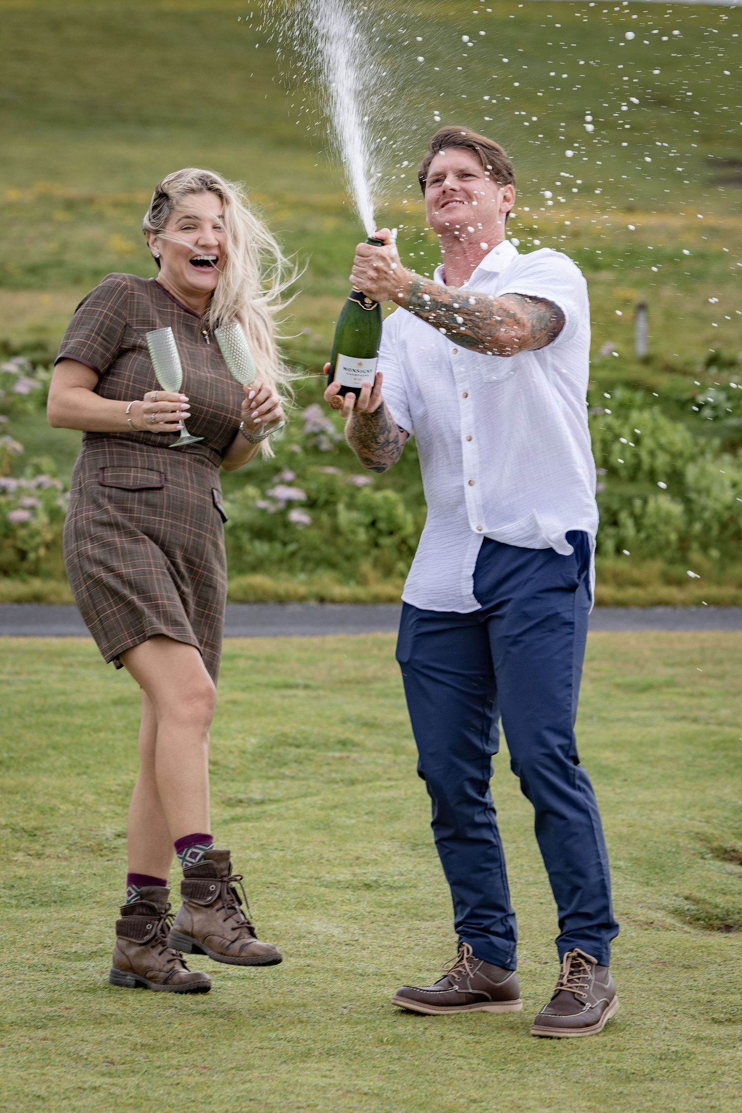 Couple celebrating outdoors as man pops champagne bottle spraying bubbly on a green lawn.