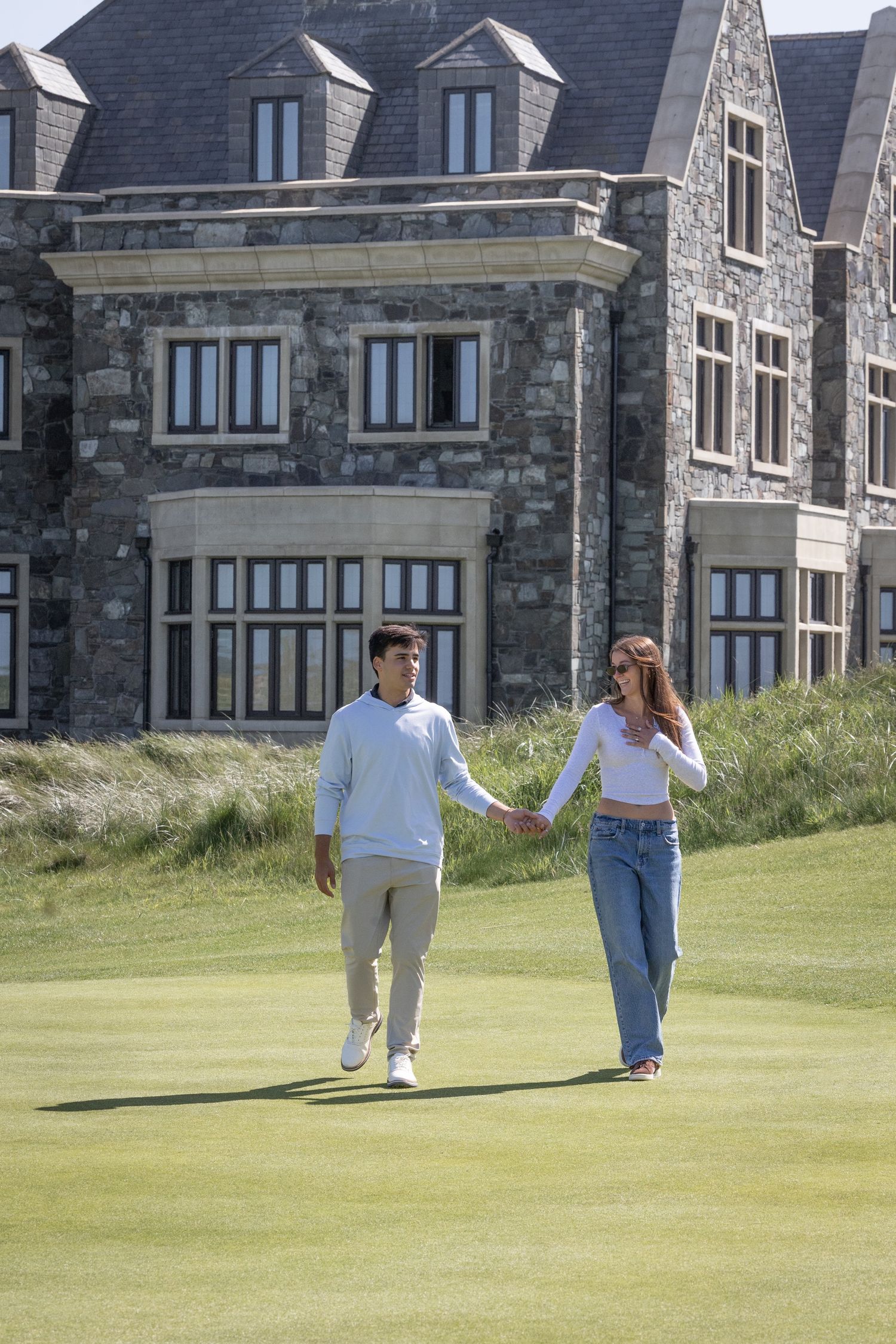 A couple holds hands while strolling across a lush green golf course beside an elegant stone manor.