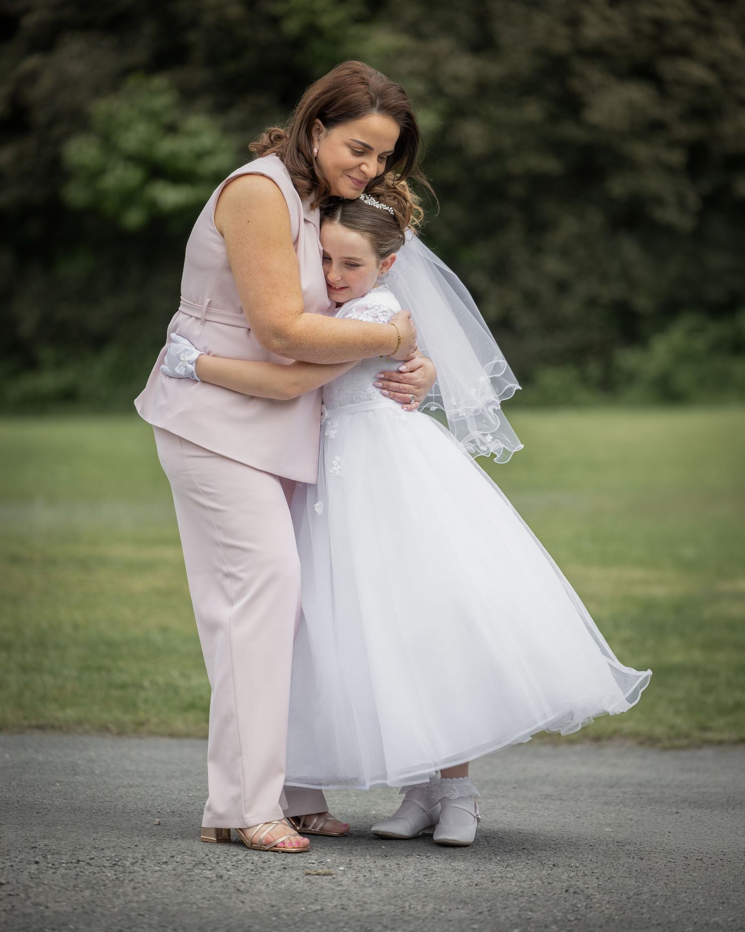 A woman in pink hugs a young girl in a white First Communion dress and veil outdoors.
