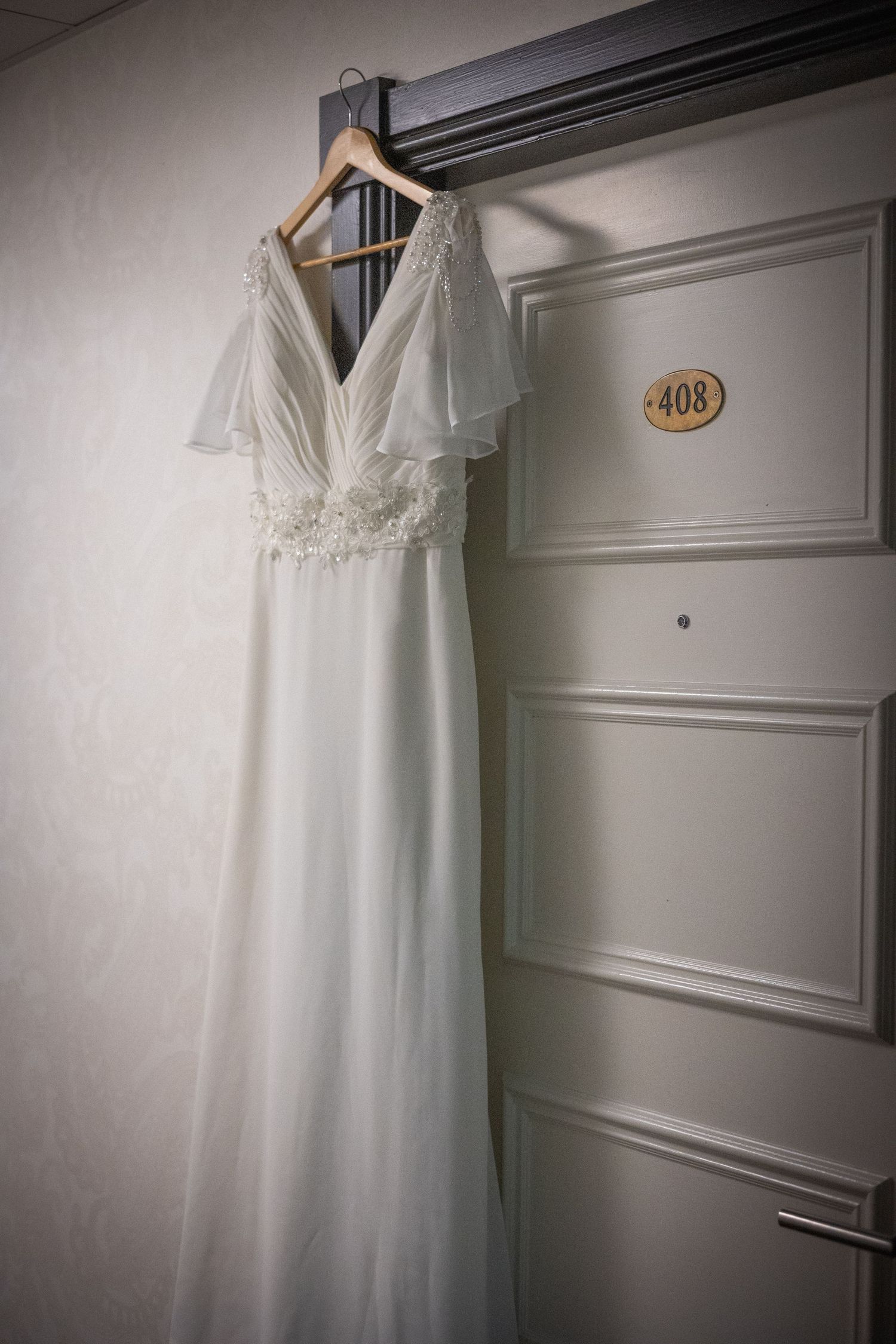 A white wedding dress hanging on a hanger against a light colored wall with room number 408 visible.
