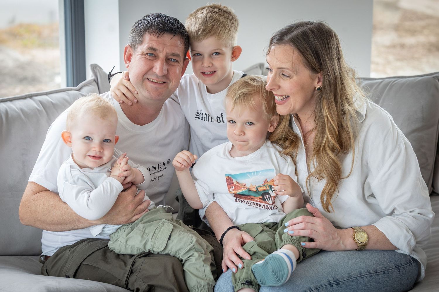 Happy family of five smiling together on a gray sofa, including parents and three young children.