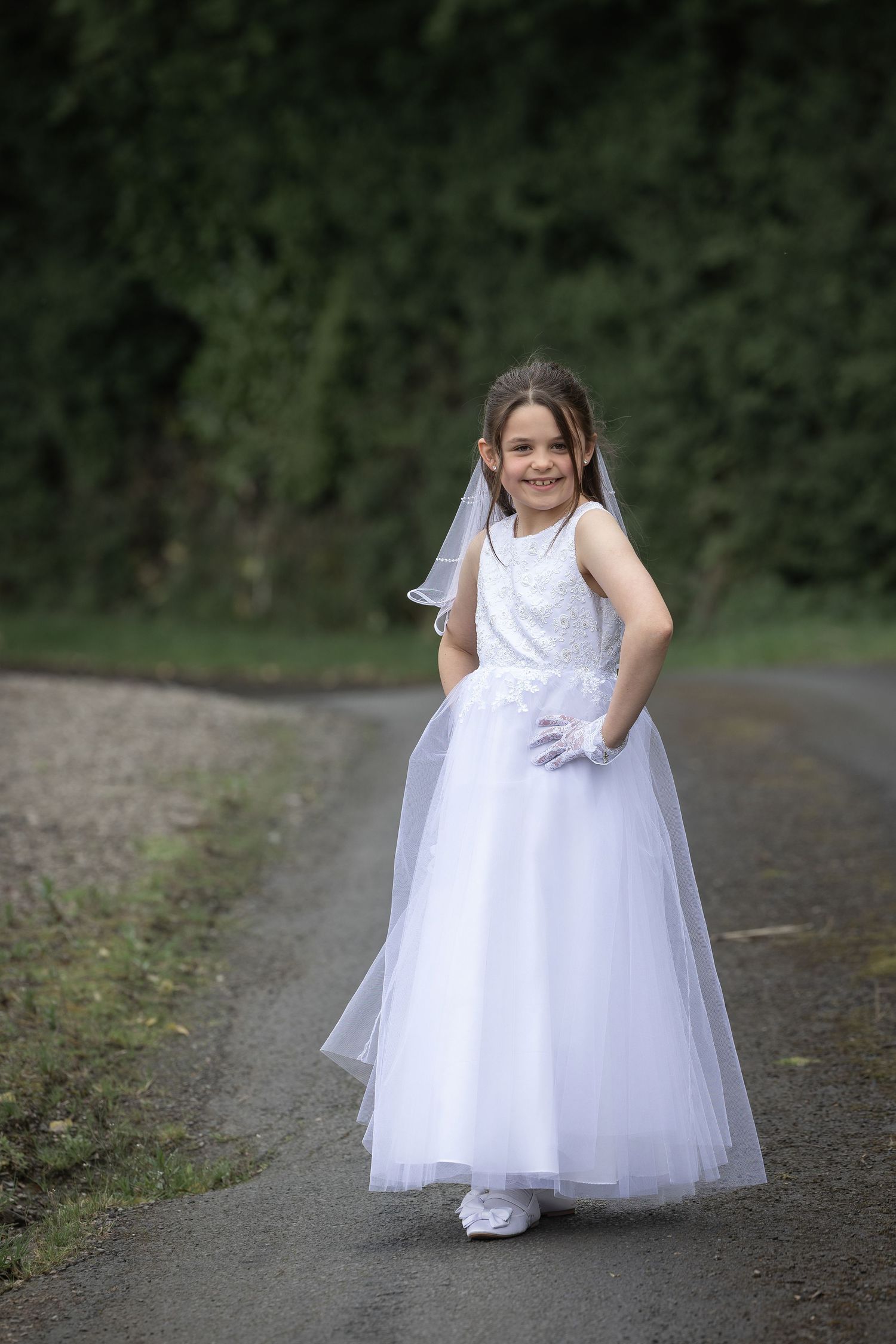 Young girl in white First Communion dress and veil smiles on a tree-lined path.