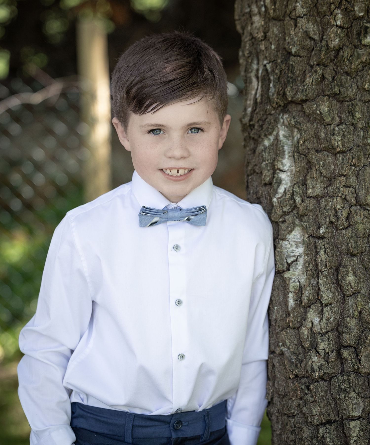 Child in formal attire poses against tree trunk wearing bow tie.