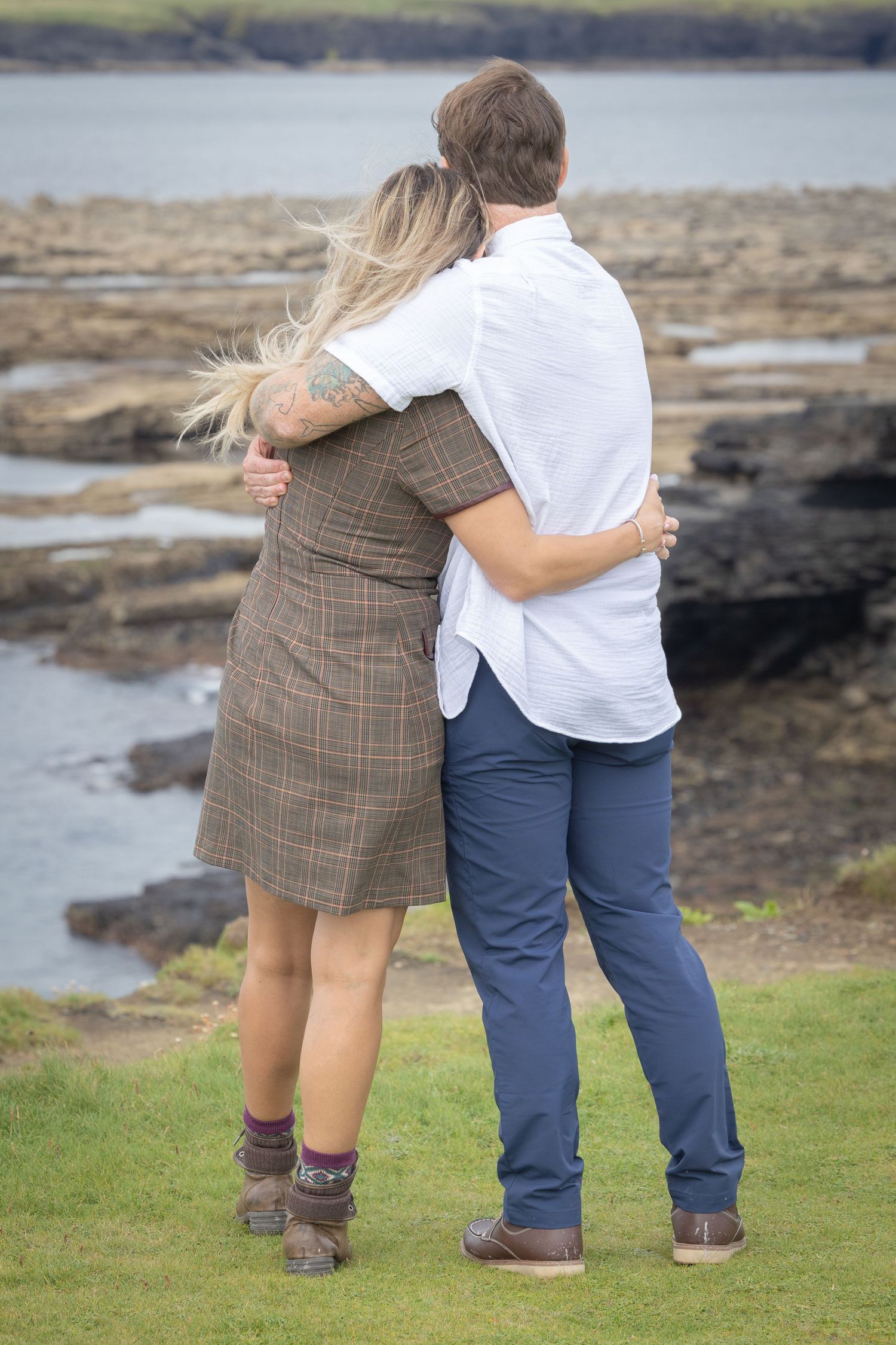 Couple embracing from behind while gazing at rocky coastal ocean view, woman with blonde hair and tattoo.